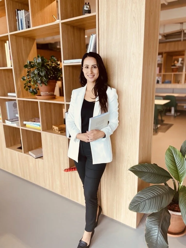 A woman with long dark hair in a white blazer and black outfit standing and smiling in an office or library space with wooden shelves and green plants.