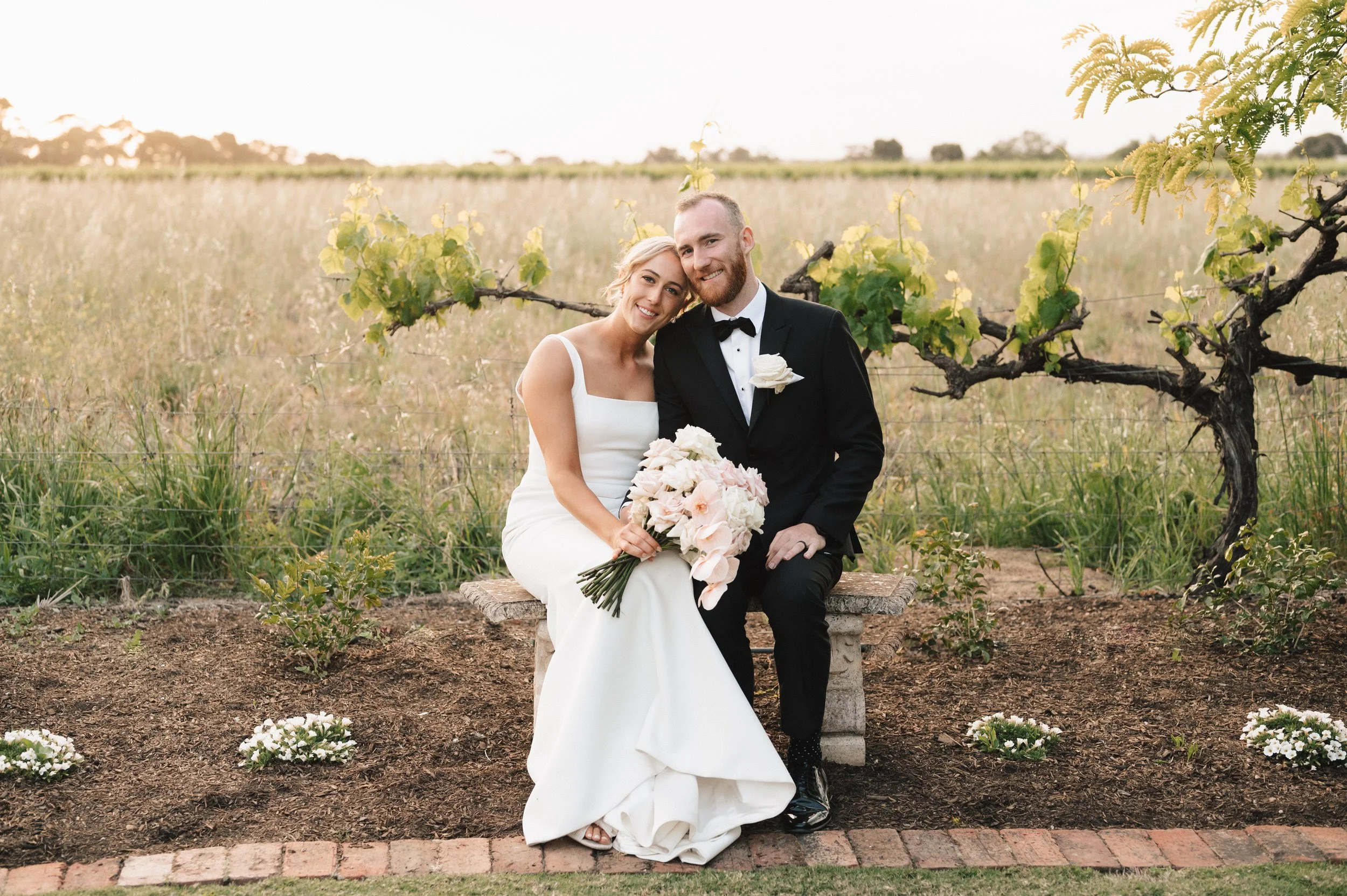 Long outdoor banquet table decorated with white tablecloths and floral centerpieces, set up under a shaded area with trees, for an event or celebration.