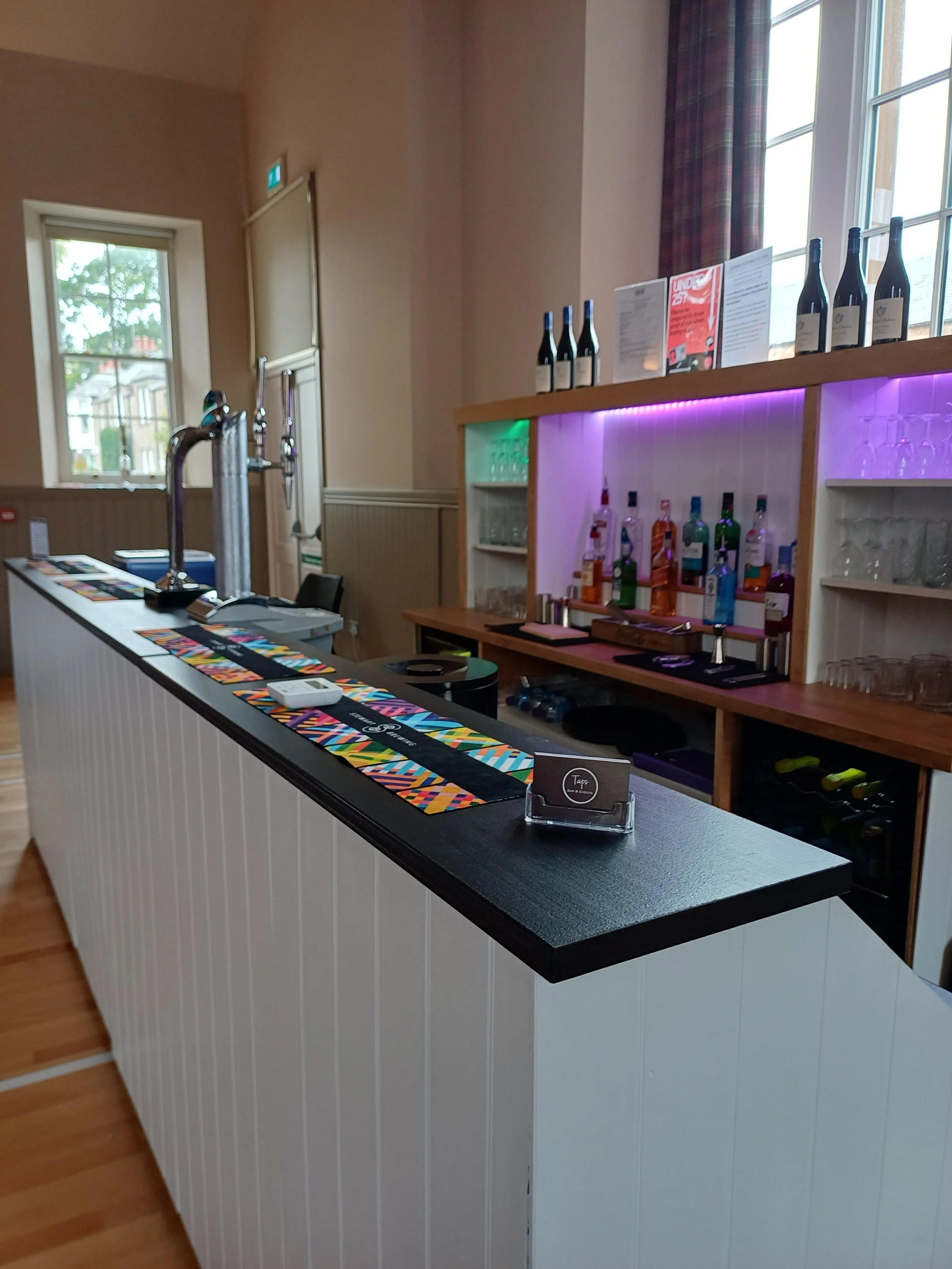Bar counter with colorful table mats, taps, and a variety of bottles and glasses, illuminated by purple and green lighting.