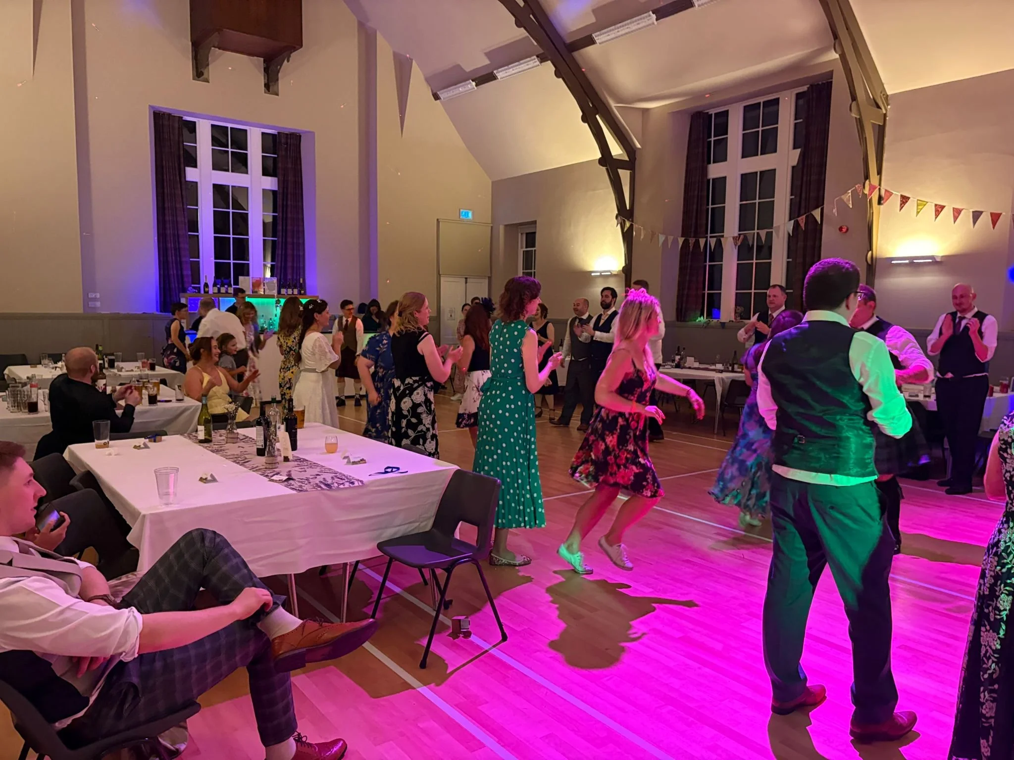 People dancing and socializing in a decorated event hall with pink and purple lighting, long tables with drinks, and bunting decorations.