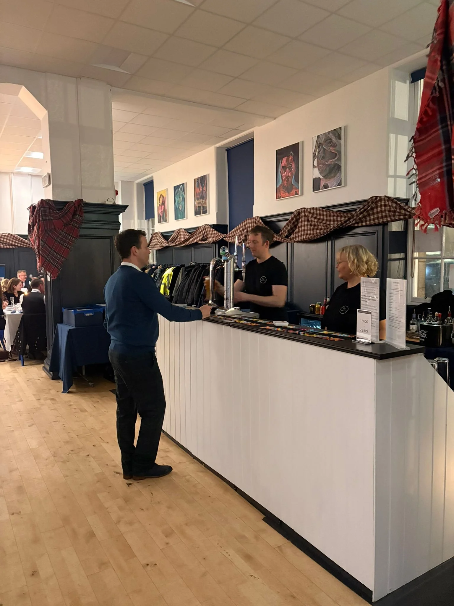 A man places an order at Taps' mobile bar at a Burns Supper event hosted at Edinburgh Academy. The bar area has a decorative checkered cloth above it, with paintings hanging on the wall behind.