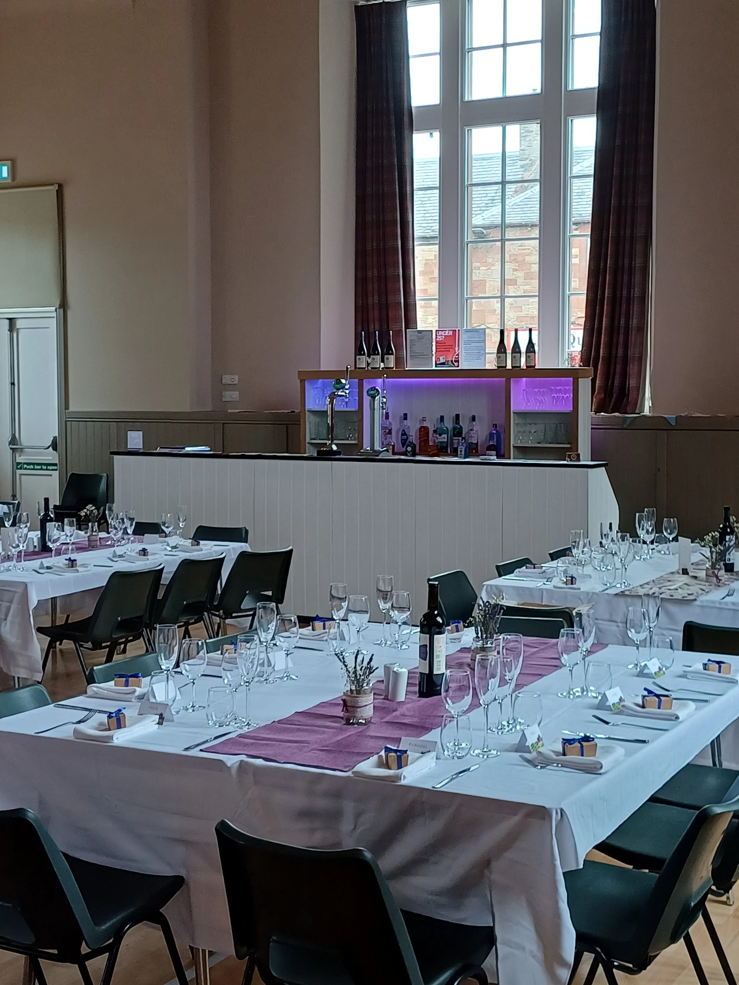 St Boswells Village hall, hosting a wedding reception. The bar from Tap Bar and Events is under the window.  Tables are set for the wedding reception, featuring white tablecloths, wine glasses, small gift boxes, and centrepieces.