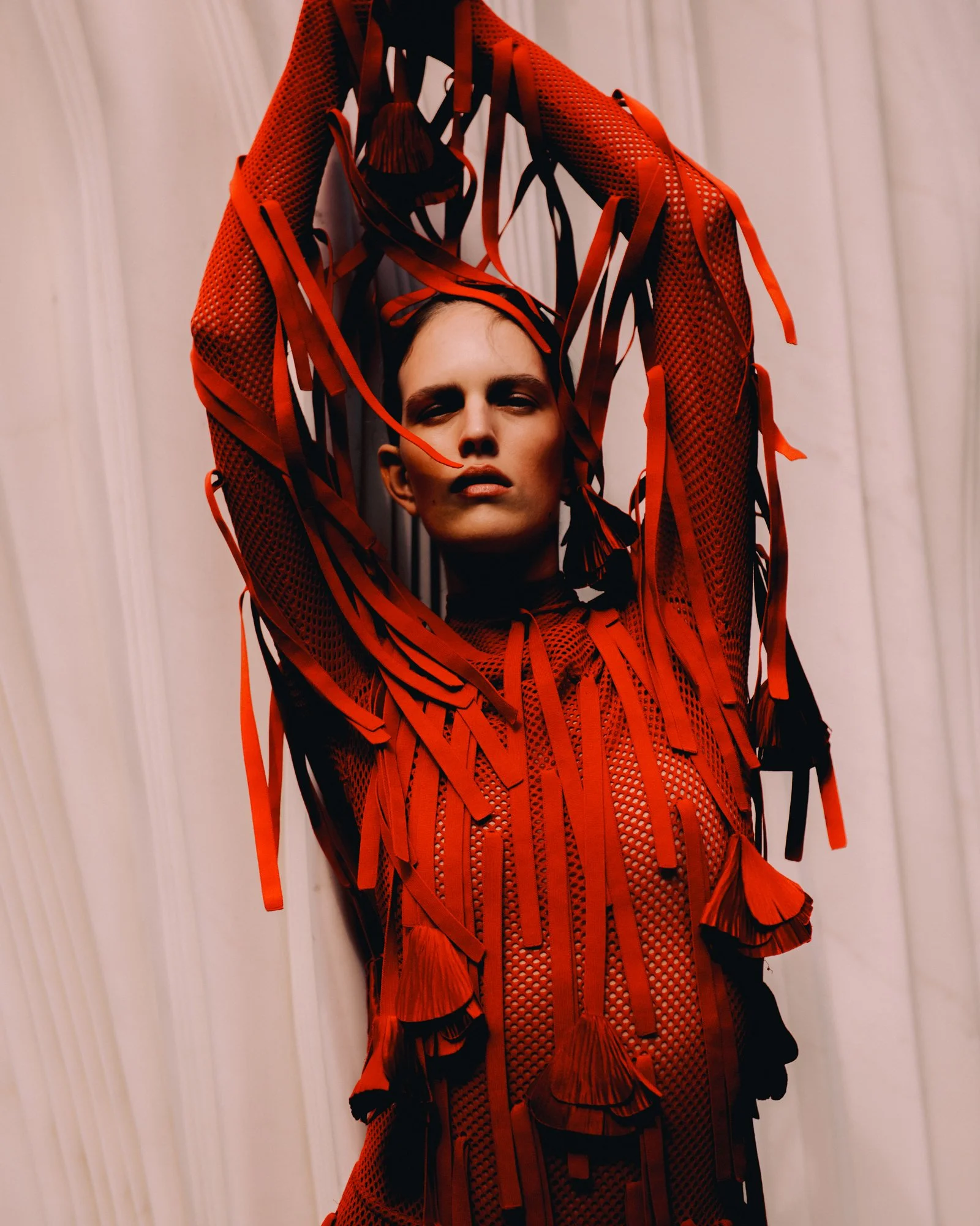 A woman wearing a red, fringed, mesh dress with long sleeves, standing against a light-colored wall, with arms raised above her head.