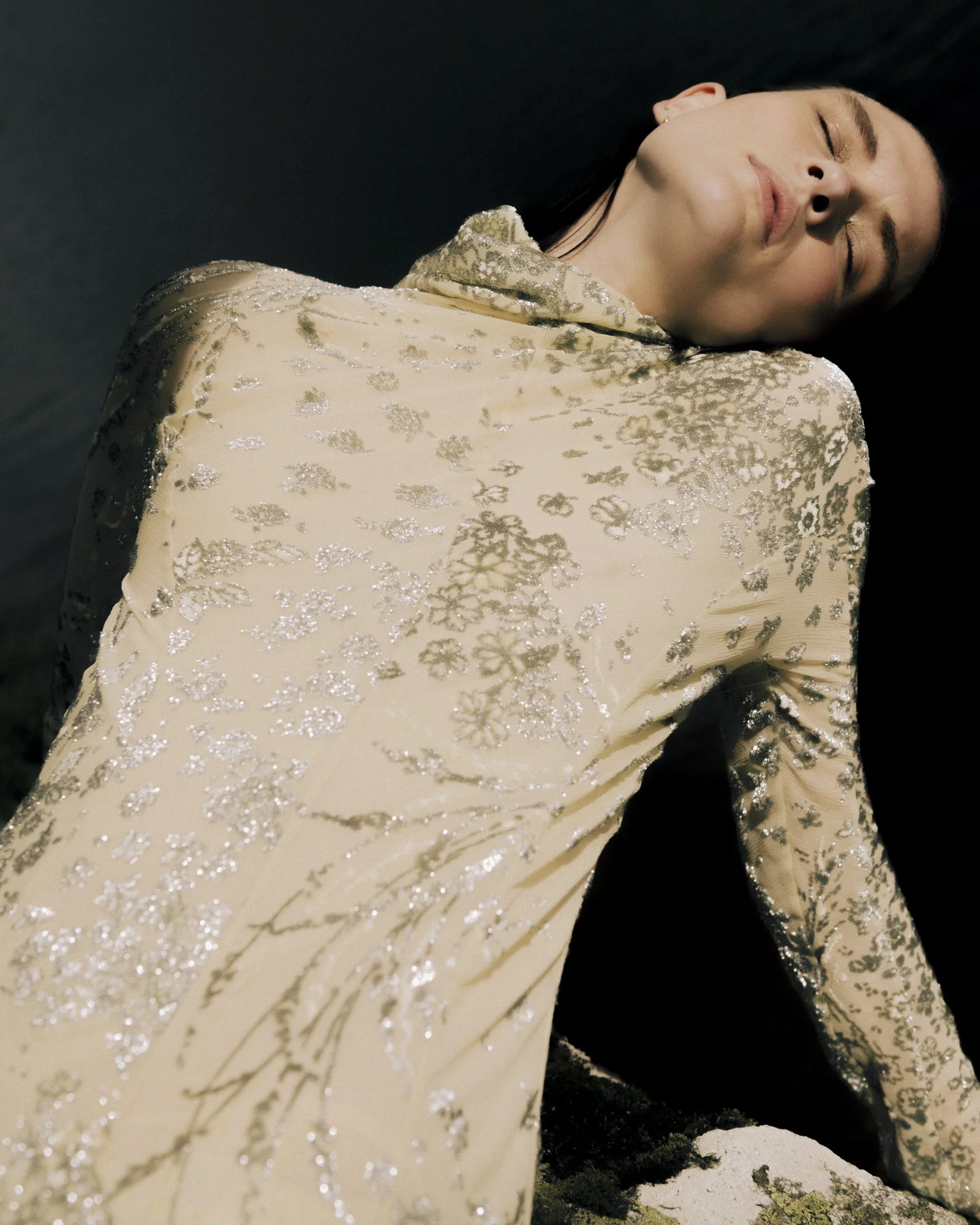 A woman with fair skin and dark hair, wearing a beige dress with silver floral patterns, is lying on her side with her eyes closed against a black background.