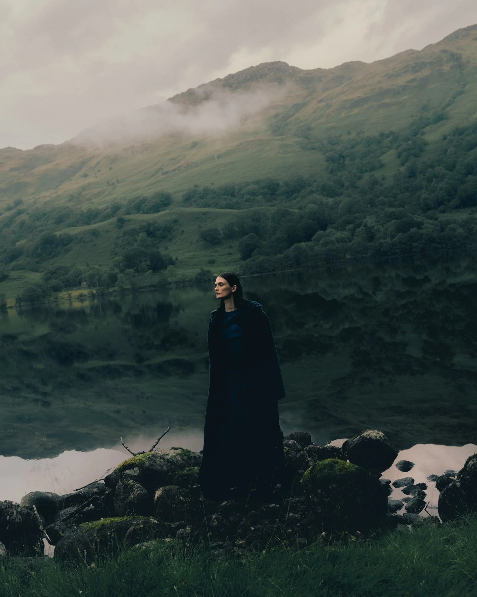 A woman in dark clothing standing on rocks beside a lake with a mountainous landscape in the background.
