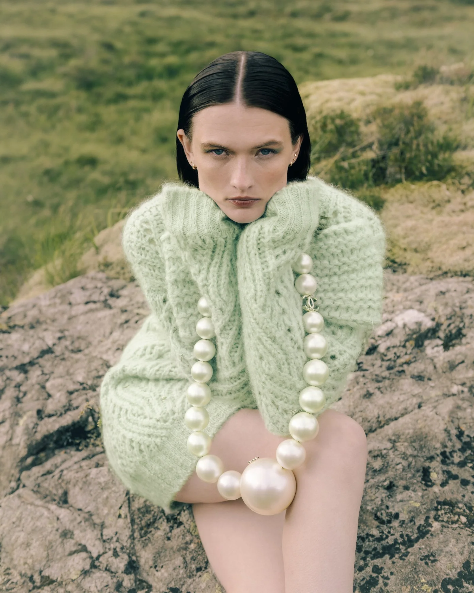 A woman with dark, slicked-back hair, wearing a light green knit sweater with pearl embellishments, sitting on rocks outdoors with a grassy landscape in the background.