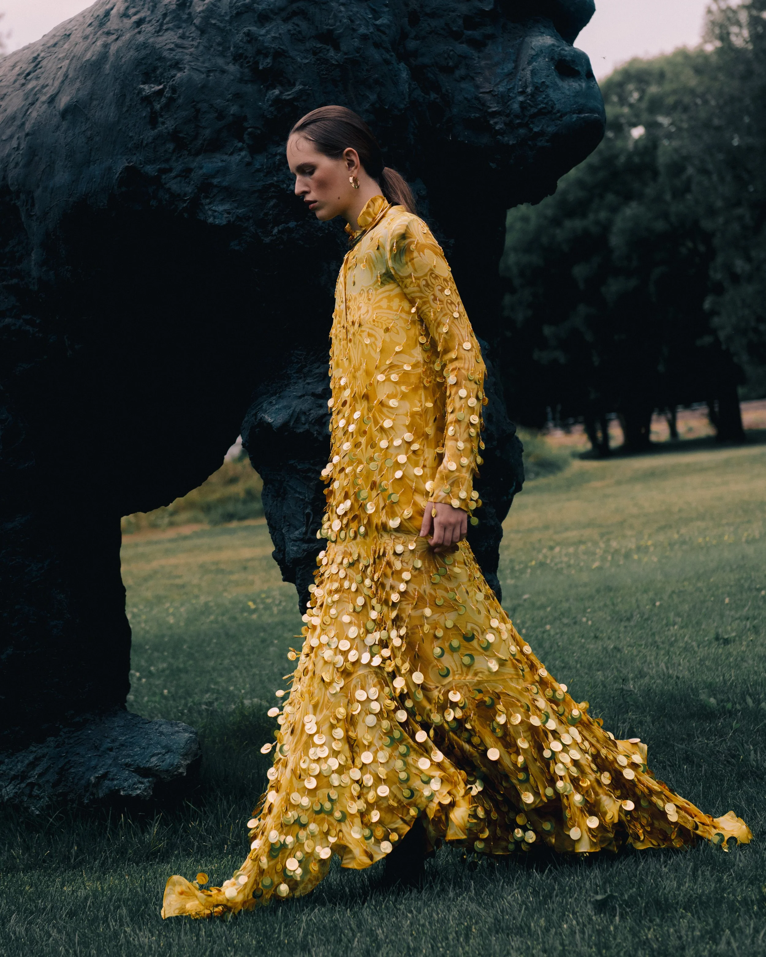 A woman in a long, yellow dress decorated with gold and green sequins walking outdoors near a large, dark sculpture of a bear.