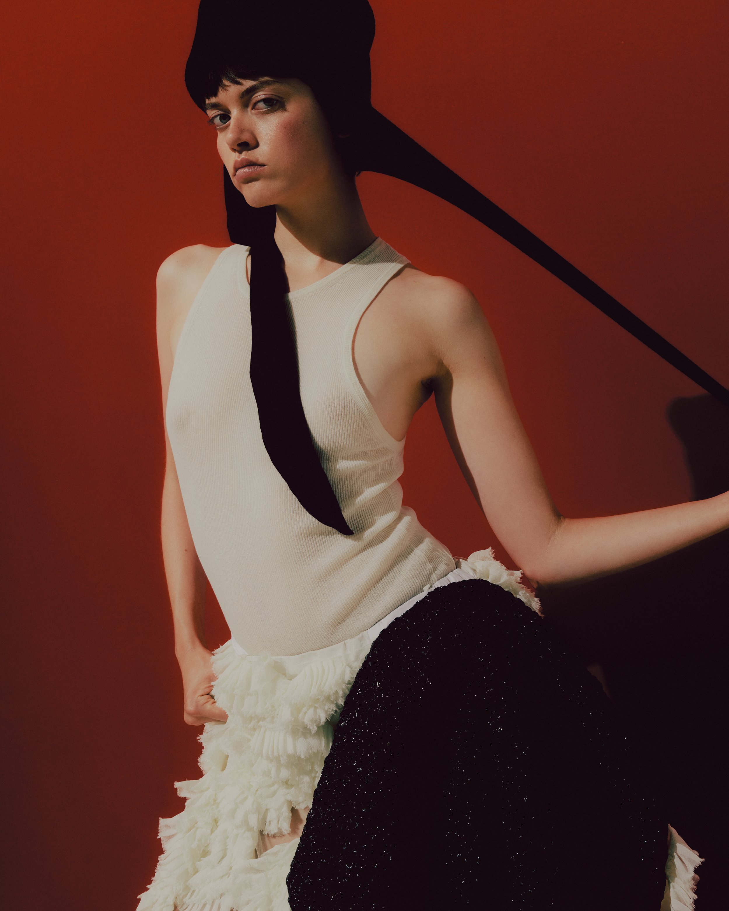 A young woman with short black hair wearing a beige sleeveless top and a layered black and white skirt against a red background.