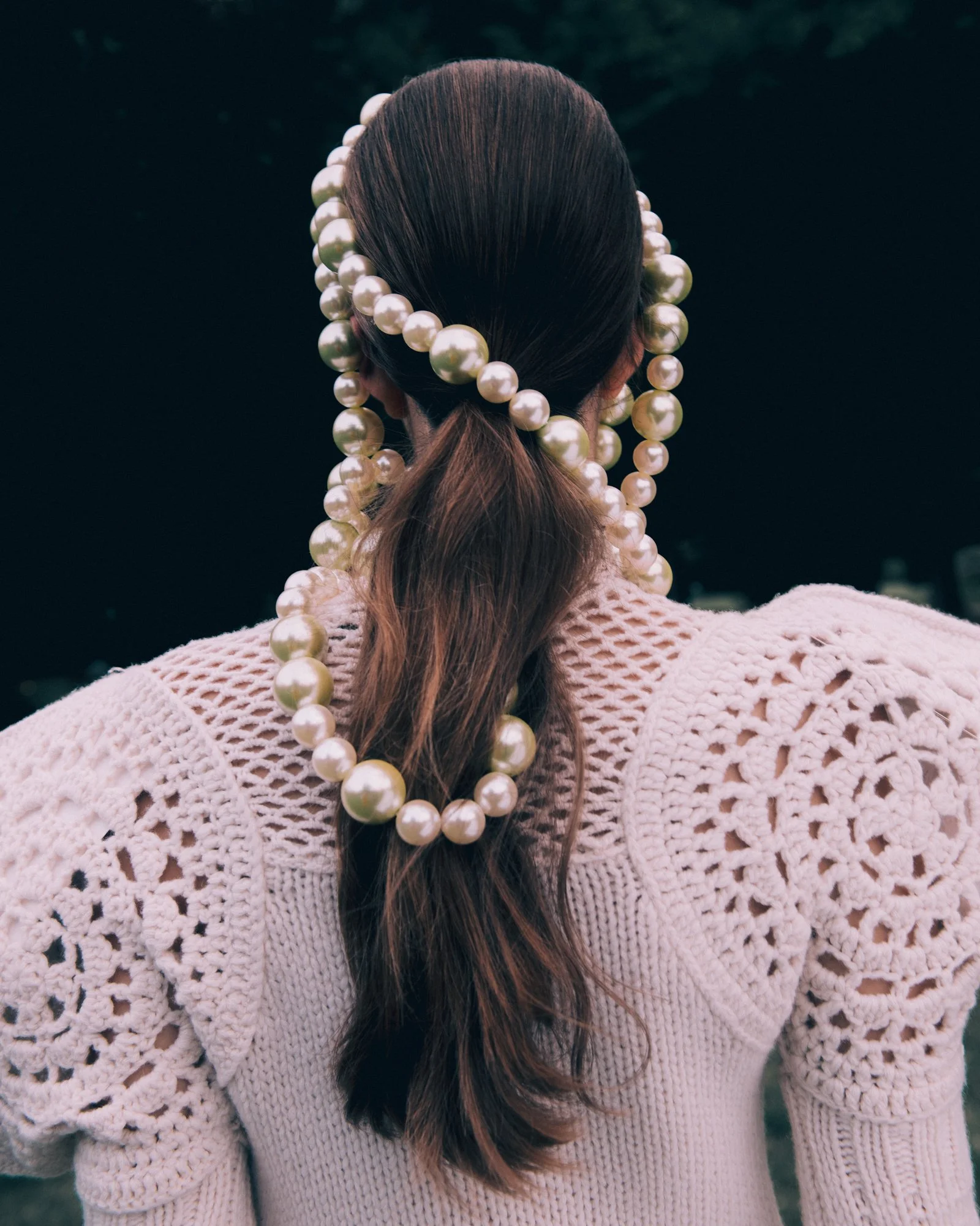 A woman with braided hair decorated with multiple pearl necklaces, wearing a light pink crocheted sweater with detailed patterns, seen from behind, outdoors at night.