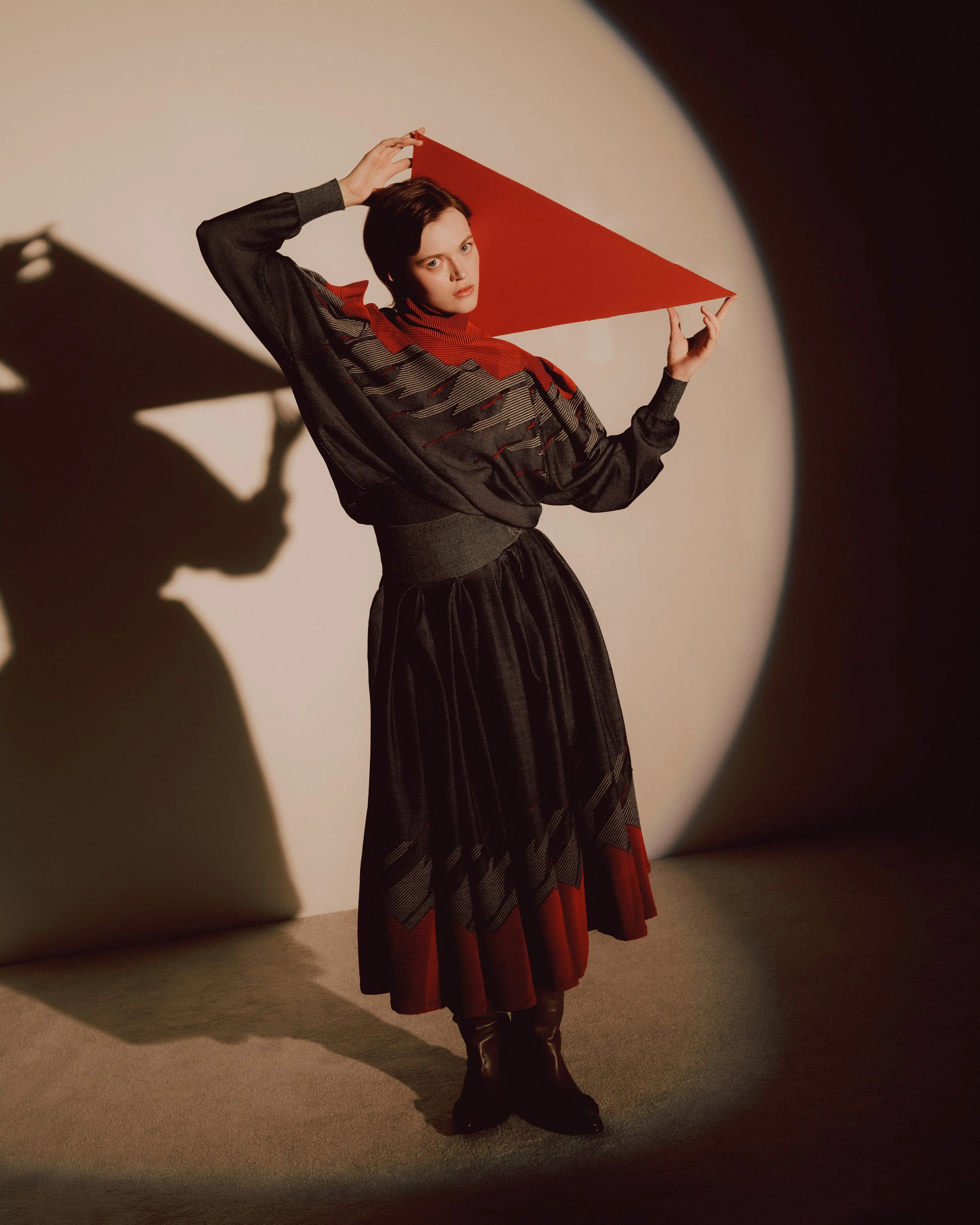 A woman wearing a dark, patterned dress with red accents, holding a red triangular object above her head, standing against a neutral background with dramatic shadows and lighting.