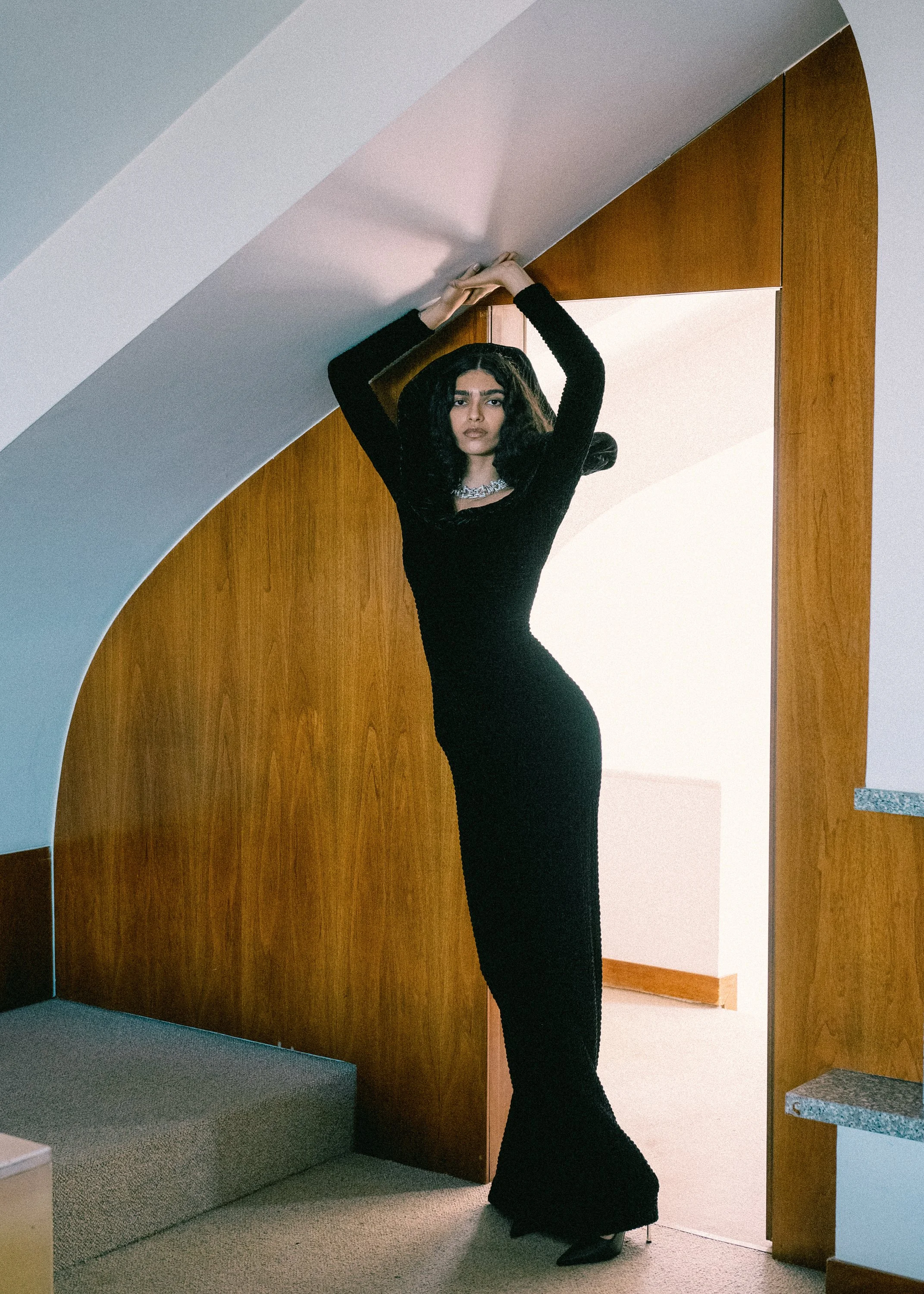A woman with long black hair wearing a form-fitting black dress and a silver necklace standing in a doorway inside a house.