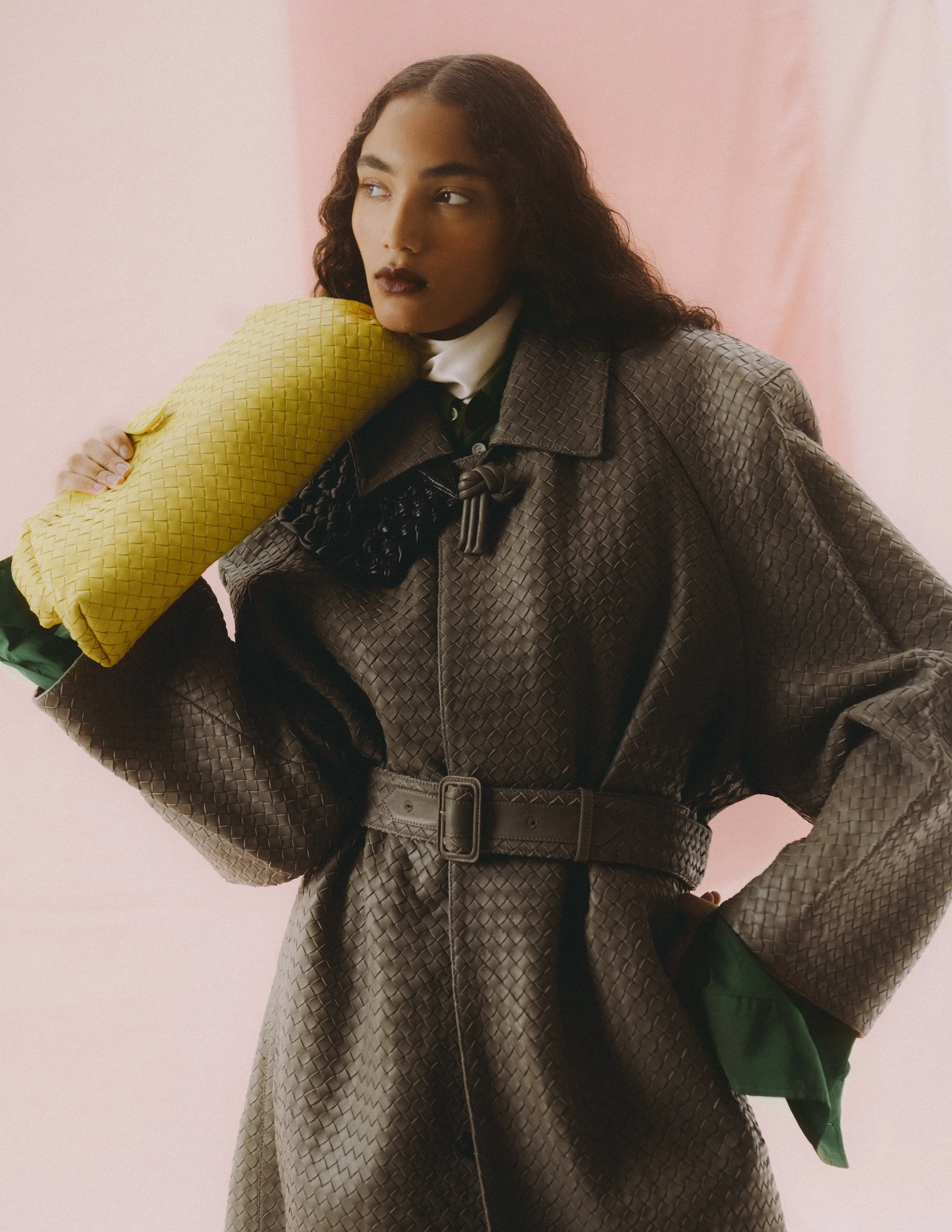 A woman with curly dark hair and dark lipstick gazes to the side, resting her head on a yellow textured pillow she holds in her hand. She wears a dark, textured, belted coat over a green shirt with a white collar, against a pink background.
