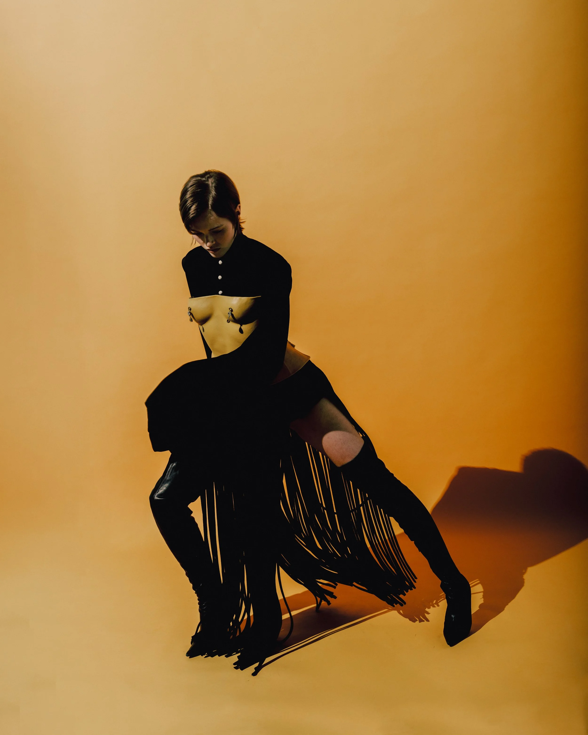 A person with short dark hair wearing a black shirt with button details is squatting in front of a yellow-orange background, with their head bowed and a shadow cast on the ground.