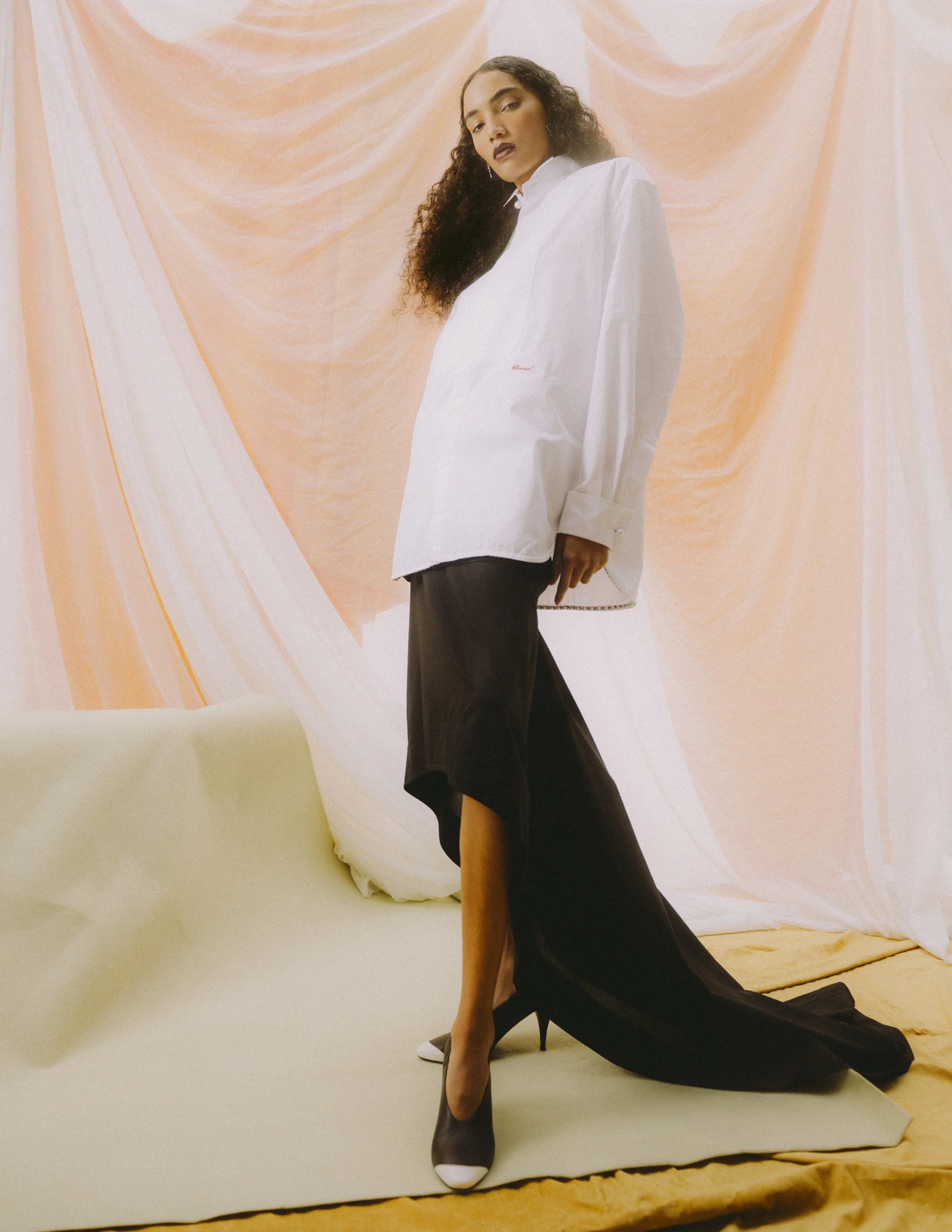 A woman with long curly hair and dark lipstick stands against a peach and white draped fabric backdrop. She's wearing a loose white button-up shirt, black layered skirt with a high slit, and pointed-toe black and white high heels. She looks confident