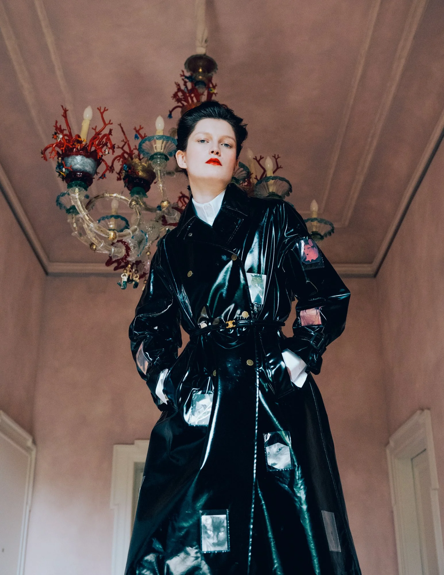 Woman with short dark hair and red lipstick wearing a black shiny trench coat with reflective patches, standing in a room with a vintage chandelier hanging from a high ceiling.