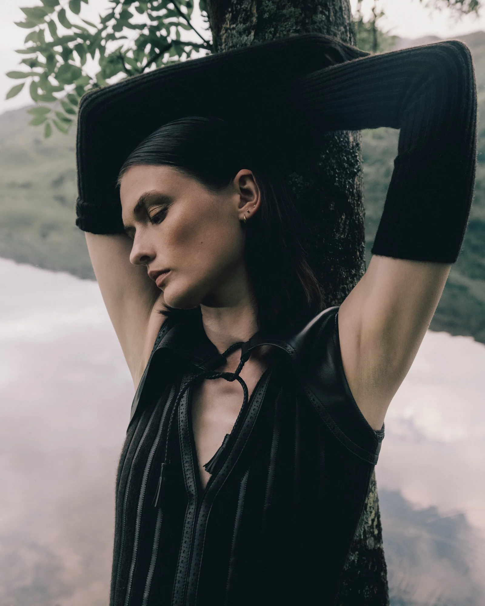 A woman with dark hair and fair skin leaning against a tree with her eyes closed, wearing a black sleeveless top and long black gloves, outdoors near a body of water.