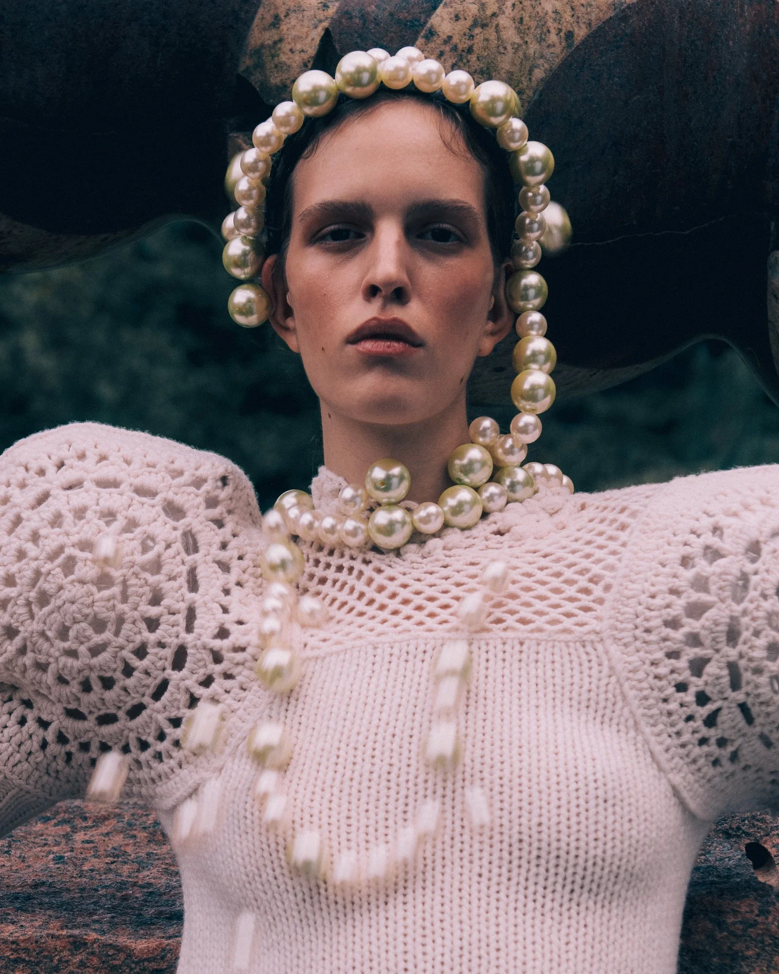 A woman with short dark hair wears a large pearl necklace and a pink crocheted sweater with puffed sleeves, standing outdoors with a green background and large rocks.