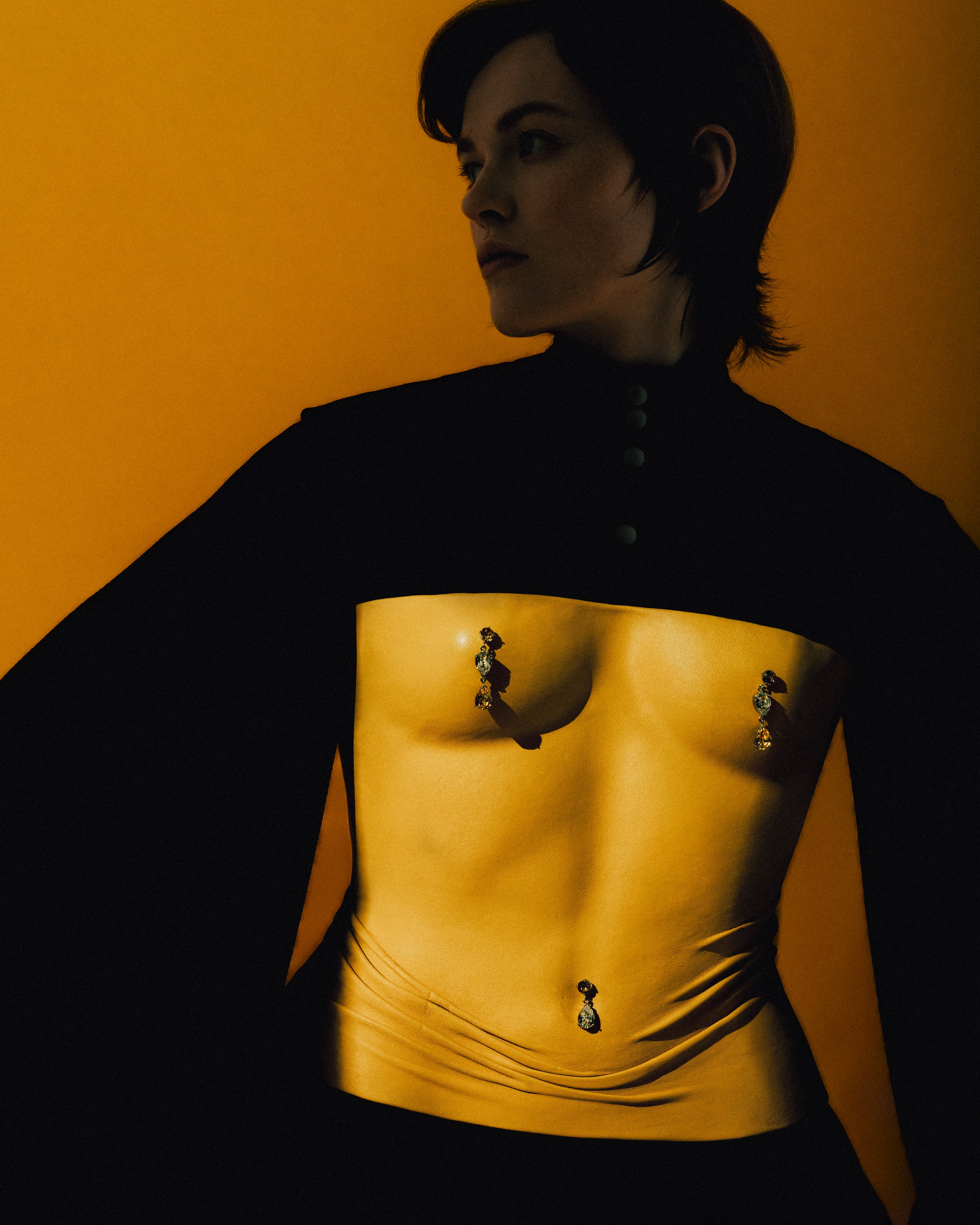A woman with short dark hair wearing a black top with long sleeves, exposing her midriff and jewelry. She is standing against a yellow background, looking to her left with a serious expression.