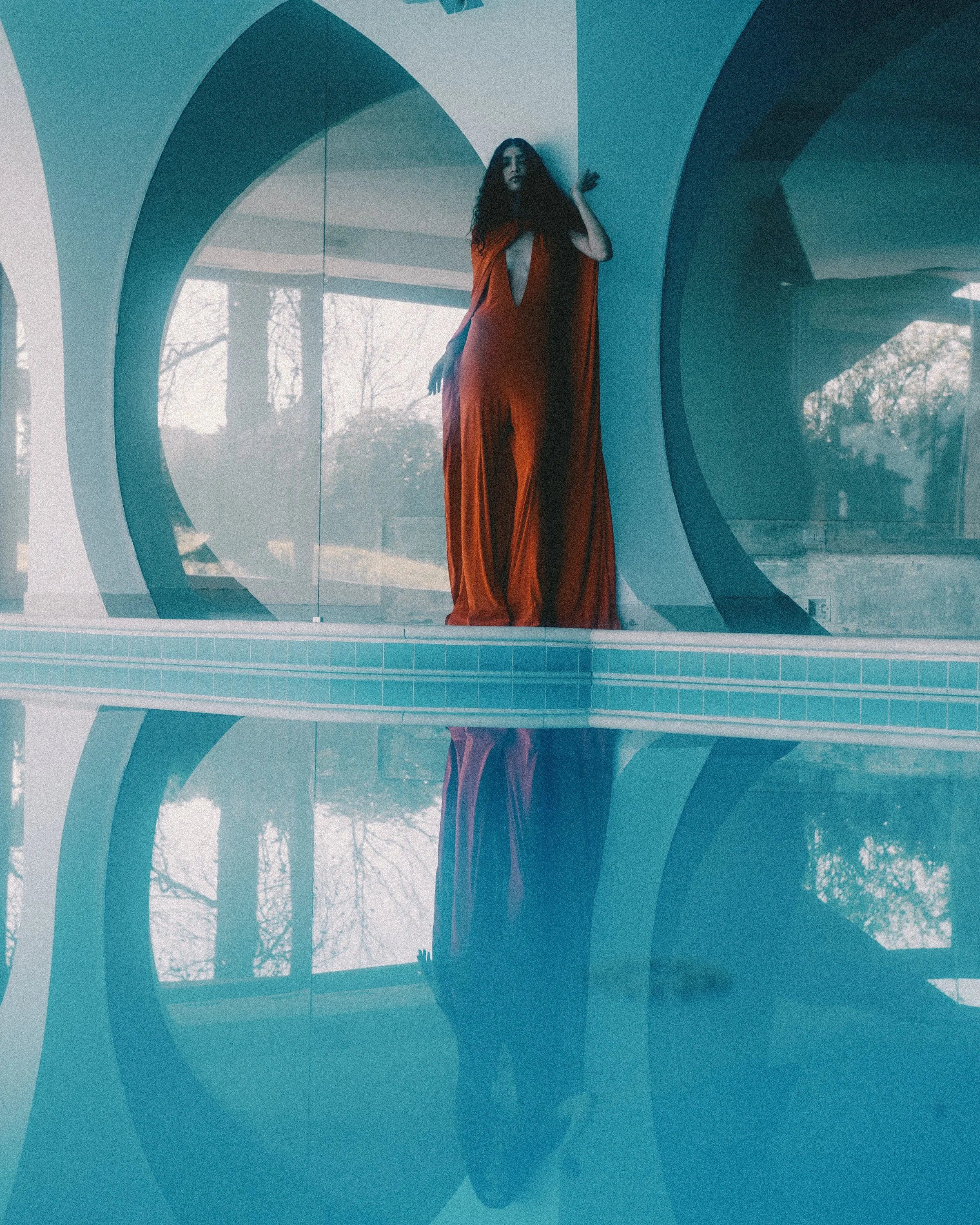 A woman in a red dress standing behind a glass window with a circular design, with her reflection visible in the water below.