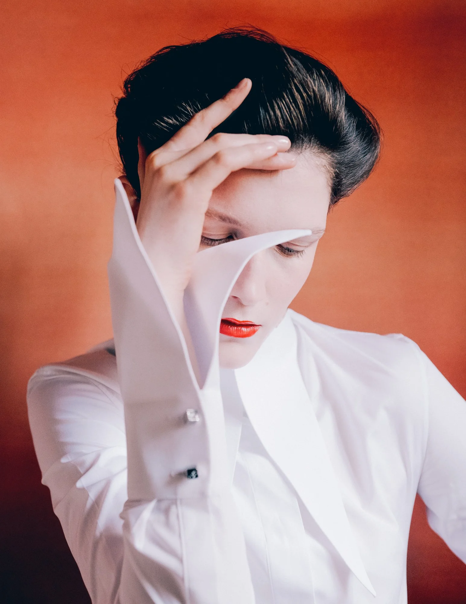 A woman with short dark hair, wearing a white shirt, has her hand on her forehead with her eyes closed, against an orange background.