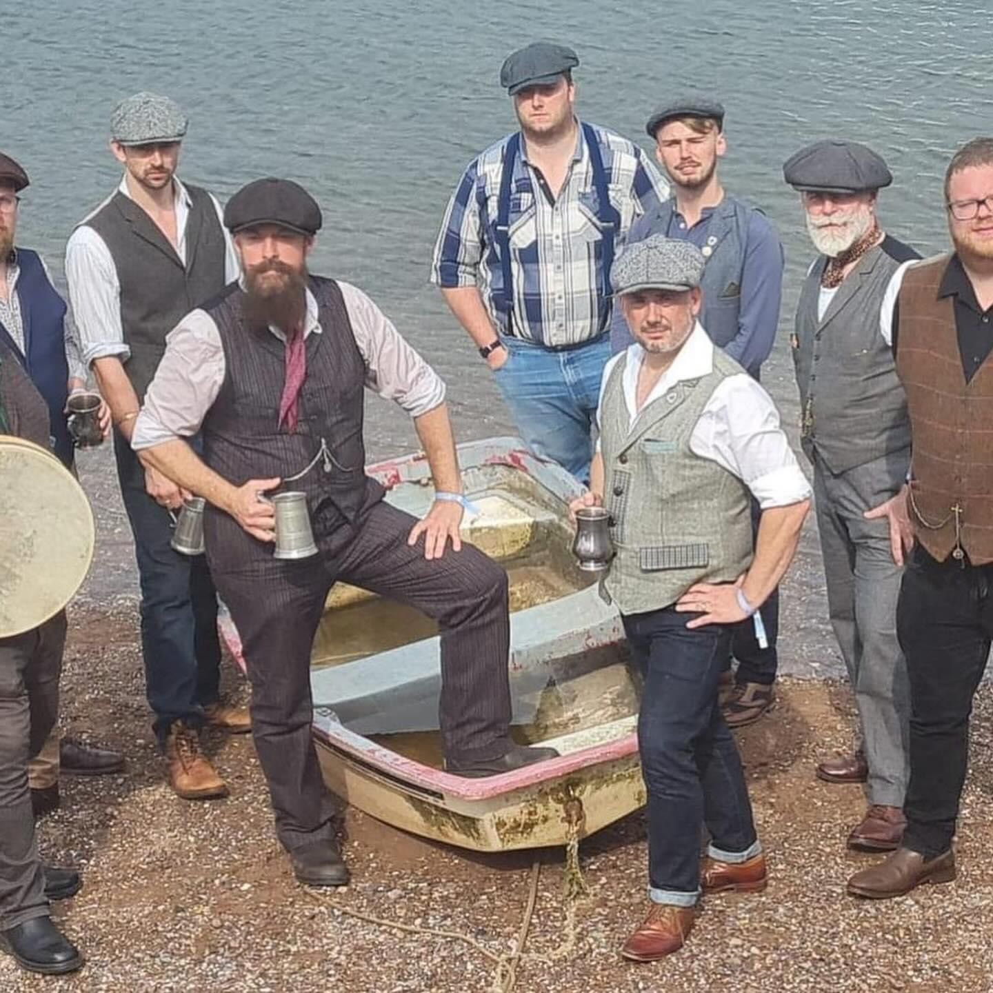 A group of men dressed in old-fashioned, vintage clothing standing around a small boat on a beach near water.