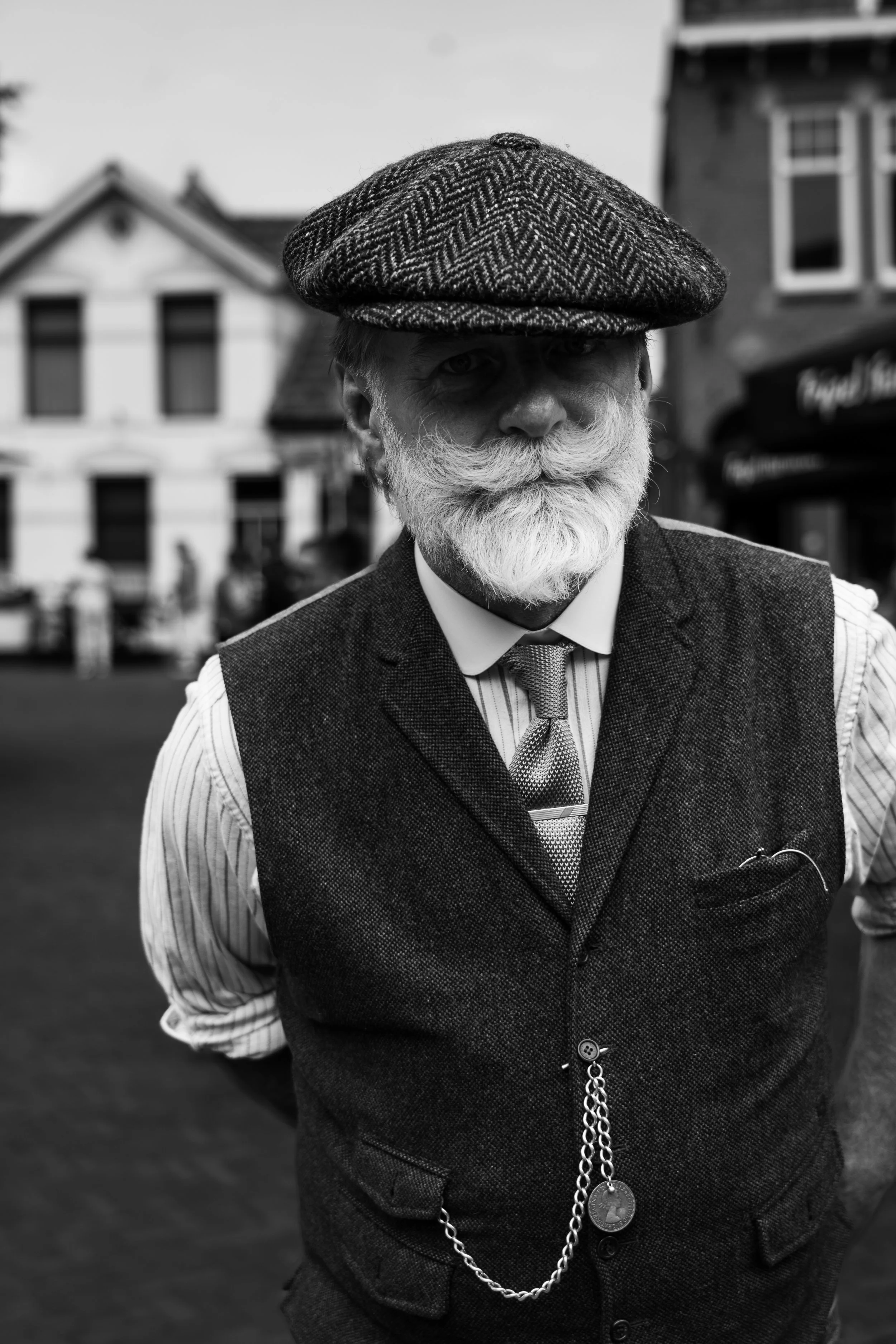 Black and white portrait of a bearded man in traditional tweed flat cap, waistcoat and tie, wearing a pocket watch chain, standing in a historic street