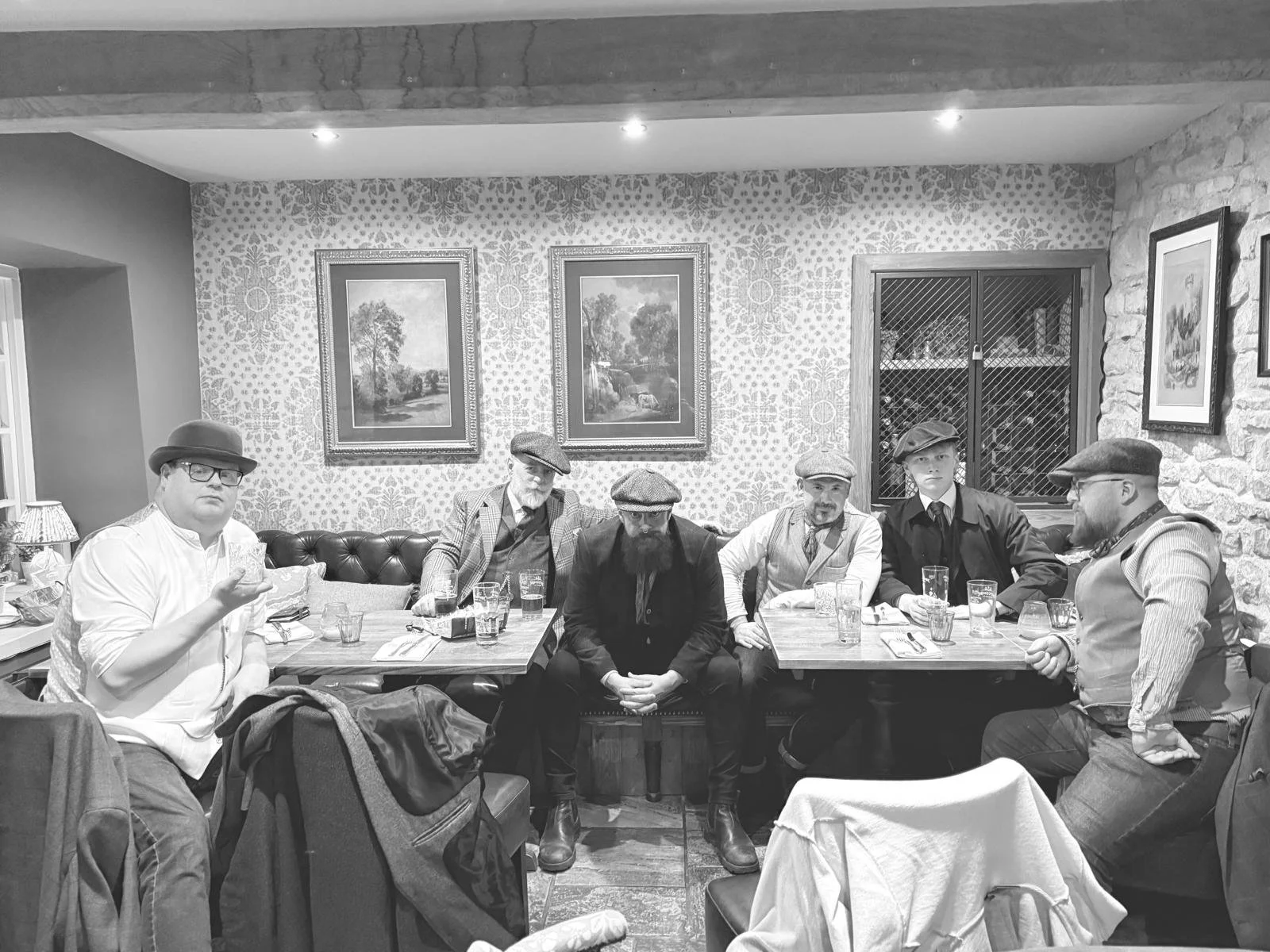 The Rusty Tubs sitting in a pub, some holding glasses. They are all wearing bakerboy-style flat caps and vintage clothing styles, with framed landscape paintings on the wallpapered walls.