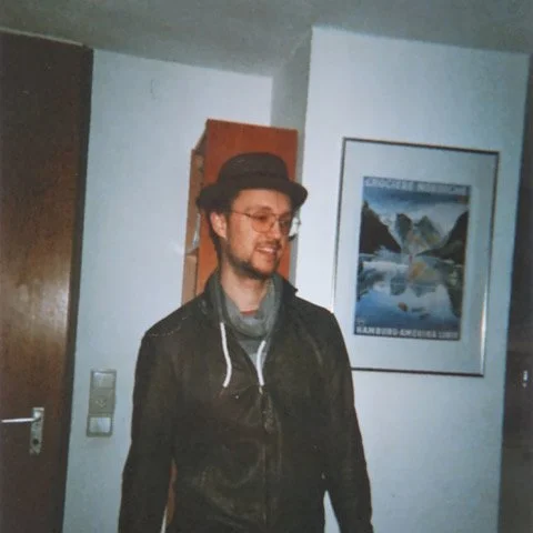 Ein Mann mit Brille und Hut steht in einem Raum. Er trägt eine Lederjacke und einen Schal. Im Hintergrund hängt ein gerahmtes Bild an der Wand.