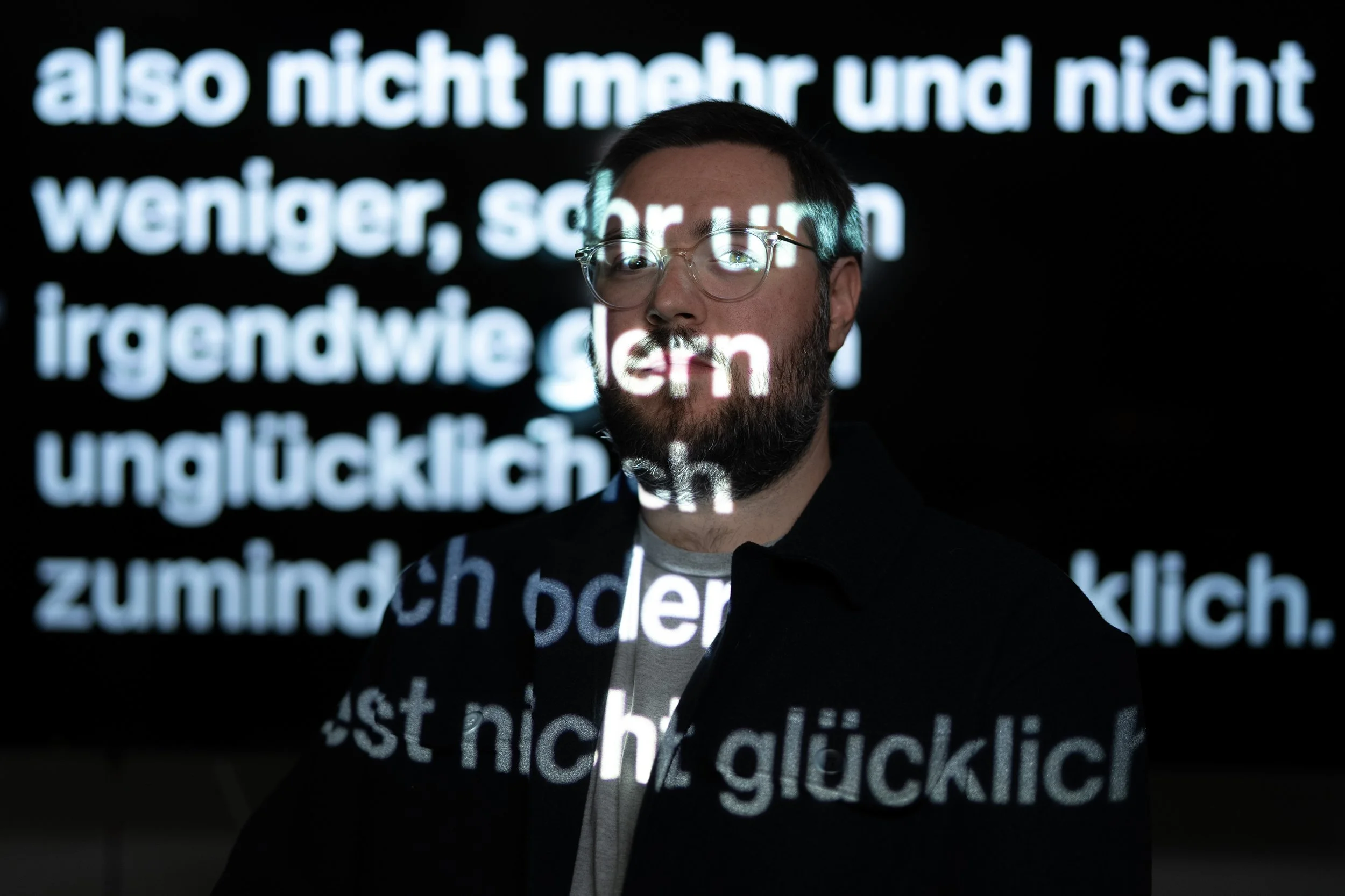 Person mit Brille und Bart vor schwarzem Hintergrund mit projiziertem Text in deutscher Sprache.