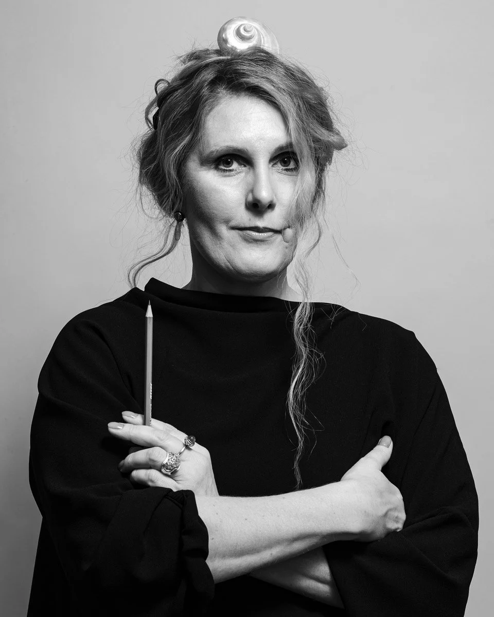 Schwarz-weiß Porträt einer Person mit lockigem Haar, einer Muschel auf dem Kopf, bekleidet mit einem schwarzen Oberteil, die einen Bleistift hält.