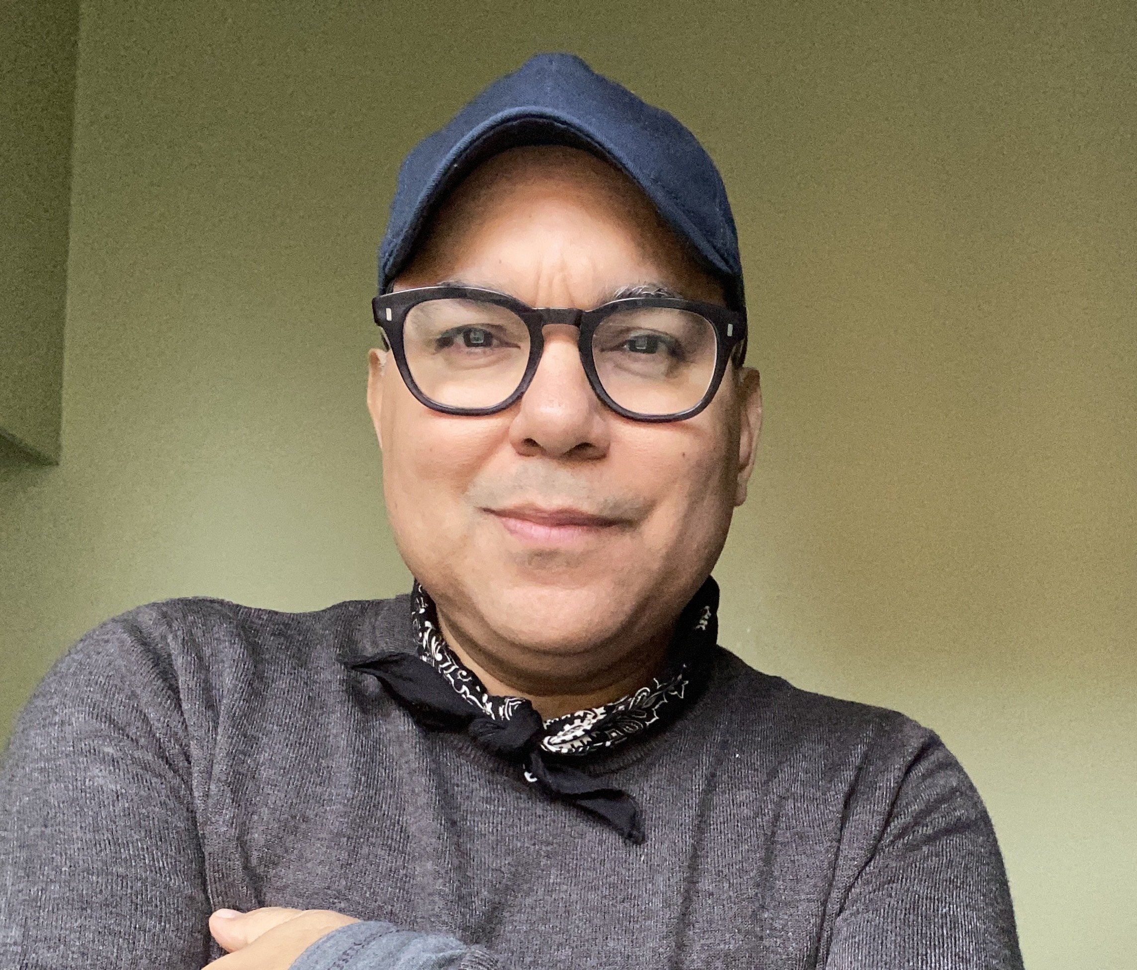 Ein Mann trägt eine dunkle Mütze, schwarze Brille und einen grauen Pullover, steht vor einer grünen Wand und lächelt leicht.