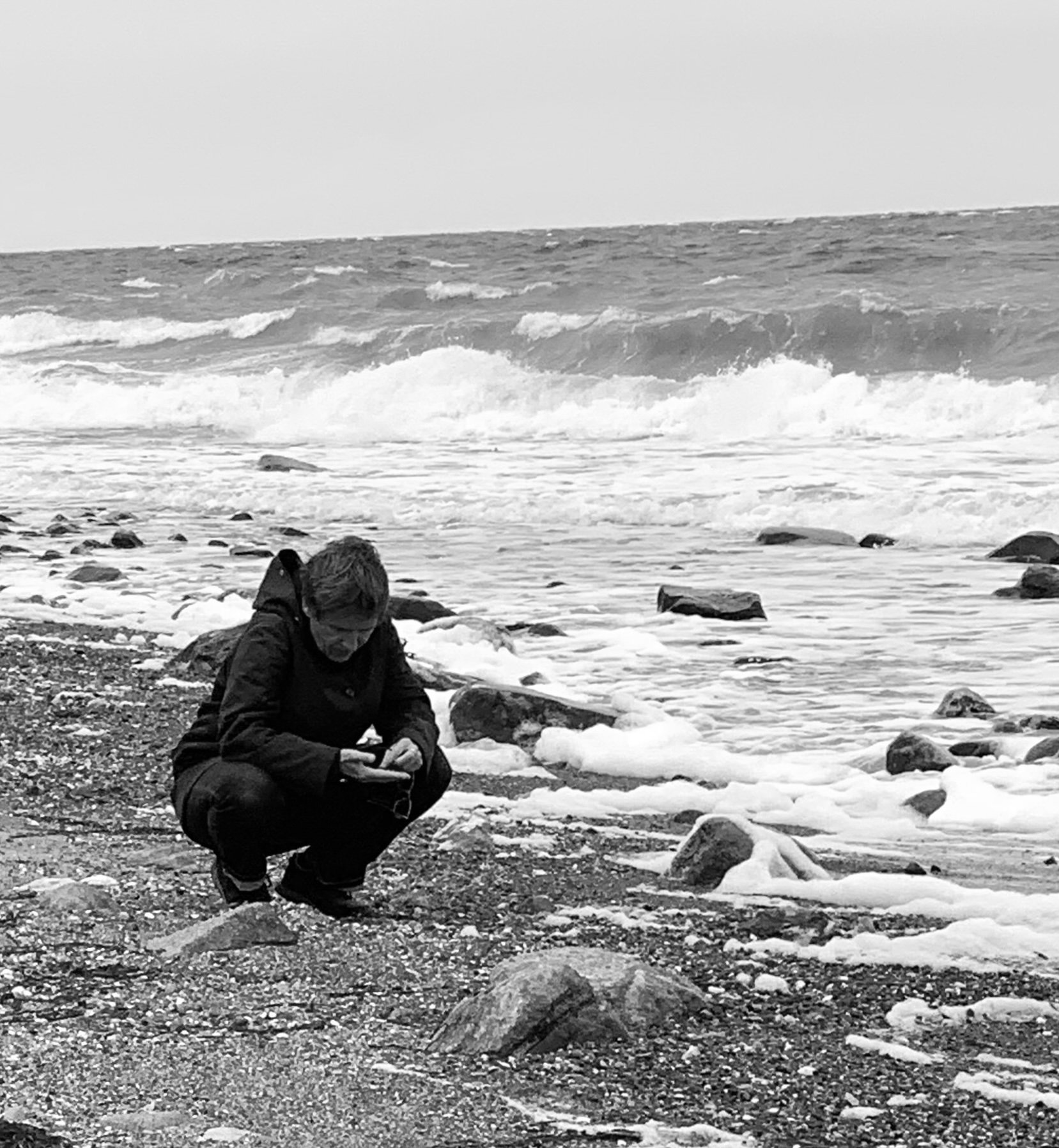 Eine Person kniet am Strand, schaut auf ihr Smartphone, mit Meereswellen im Hintergrund.