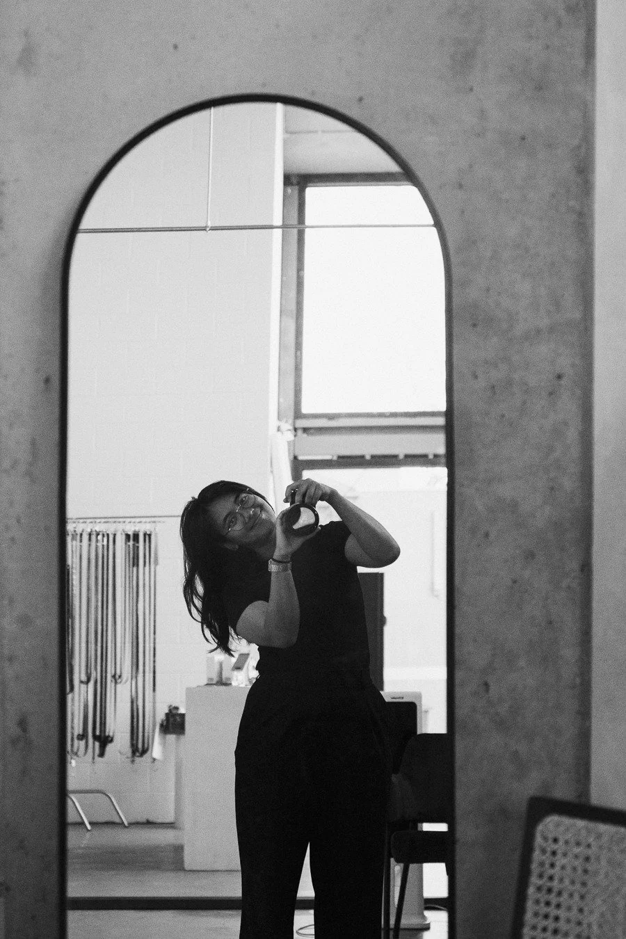 A woman taking a mirror selfie in a room with a large arched mirror, wearing glasses and black clothing.