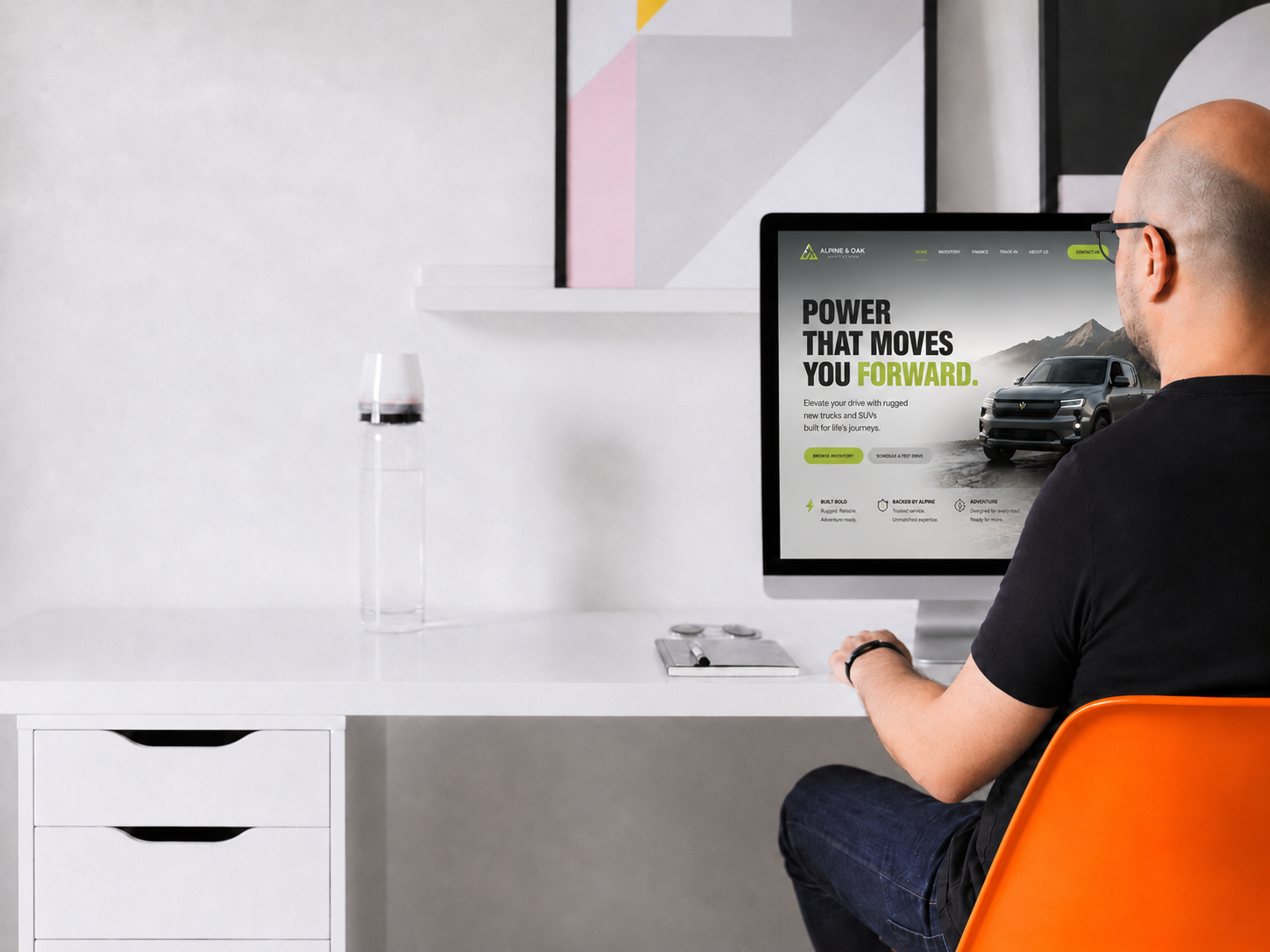 A man sitting at a desk looking at a computer monitor with a website about trucks or SUVs. The desk has a glass with red wine, a closed notebook, and a plate with coins. There is a colorful abstract painting on the wall behind him.