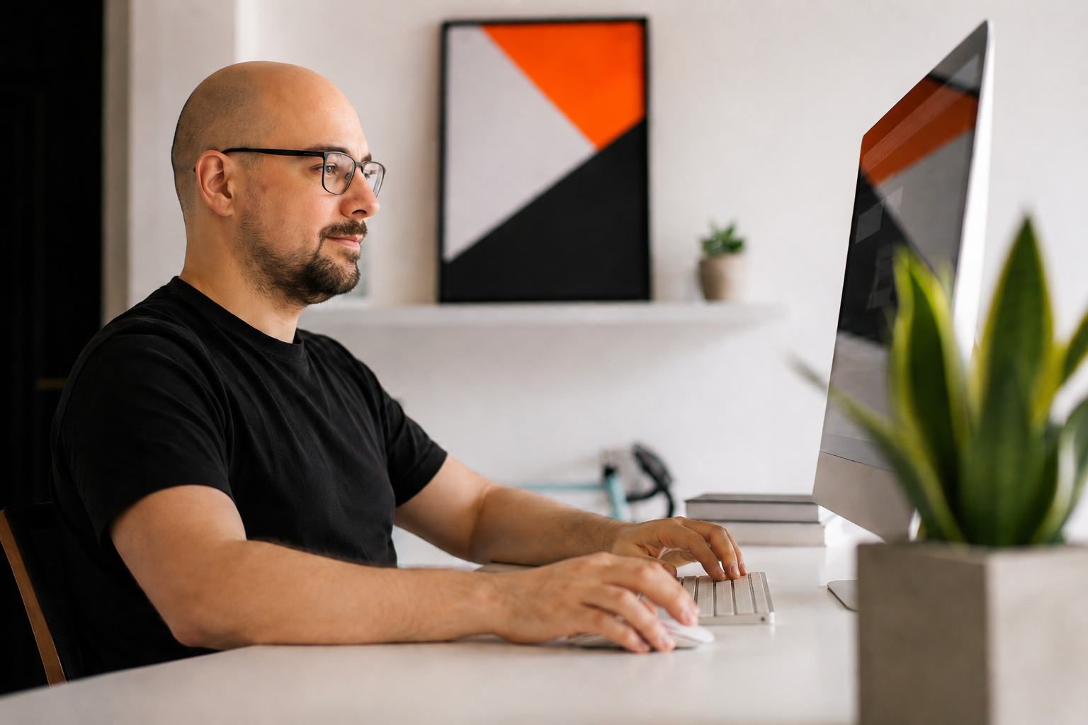 Man with glasses working on a computer at a white desk in a modern room with abstract wall art and a potted plant.