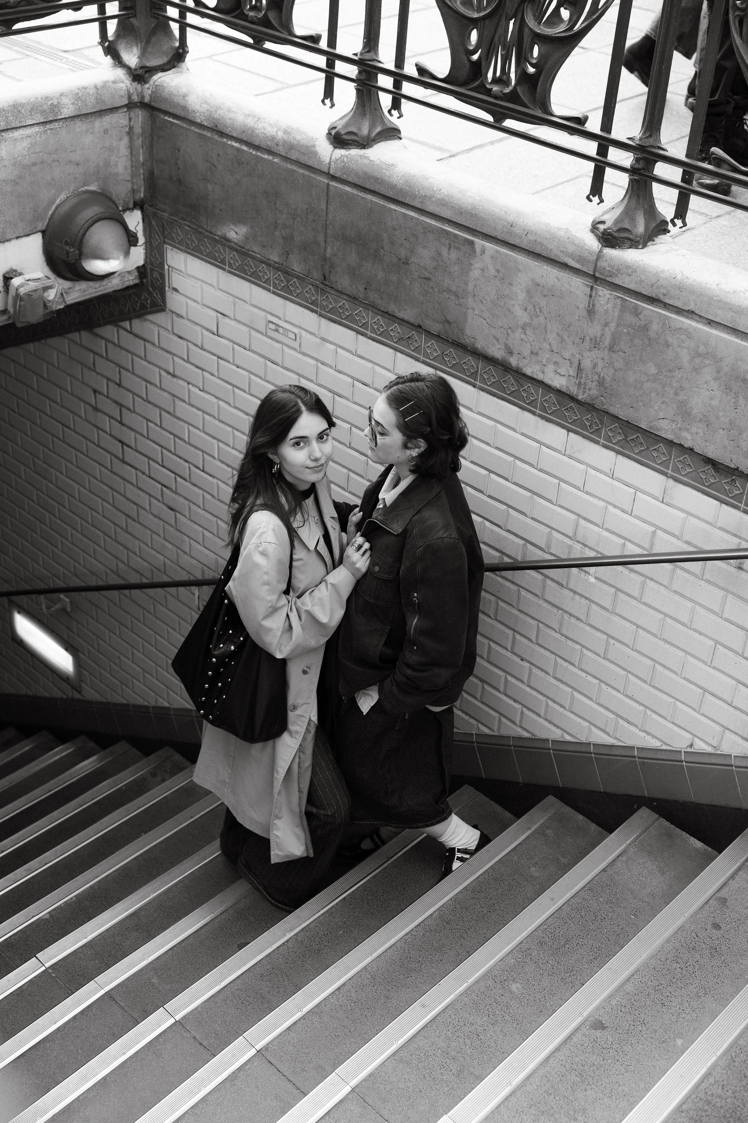 Deux jeunes femmes se tiennent sur un escalier intérieur, regardant le camera. L'une porte un trench beige avec un sac à main noir, l'autre porte une veste en jean sombre et porte des barres à cheveux dans ses cheveux.