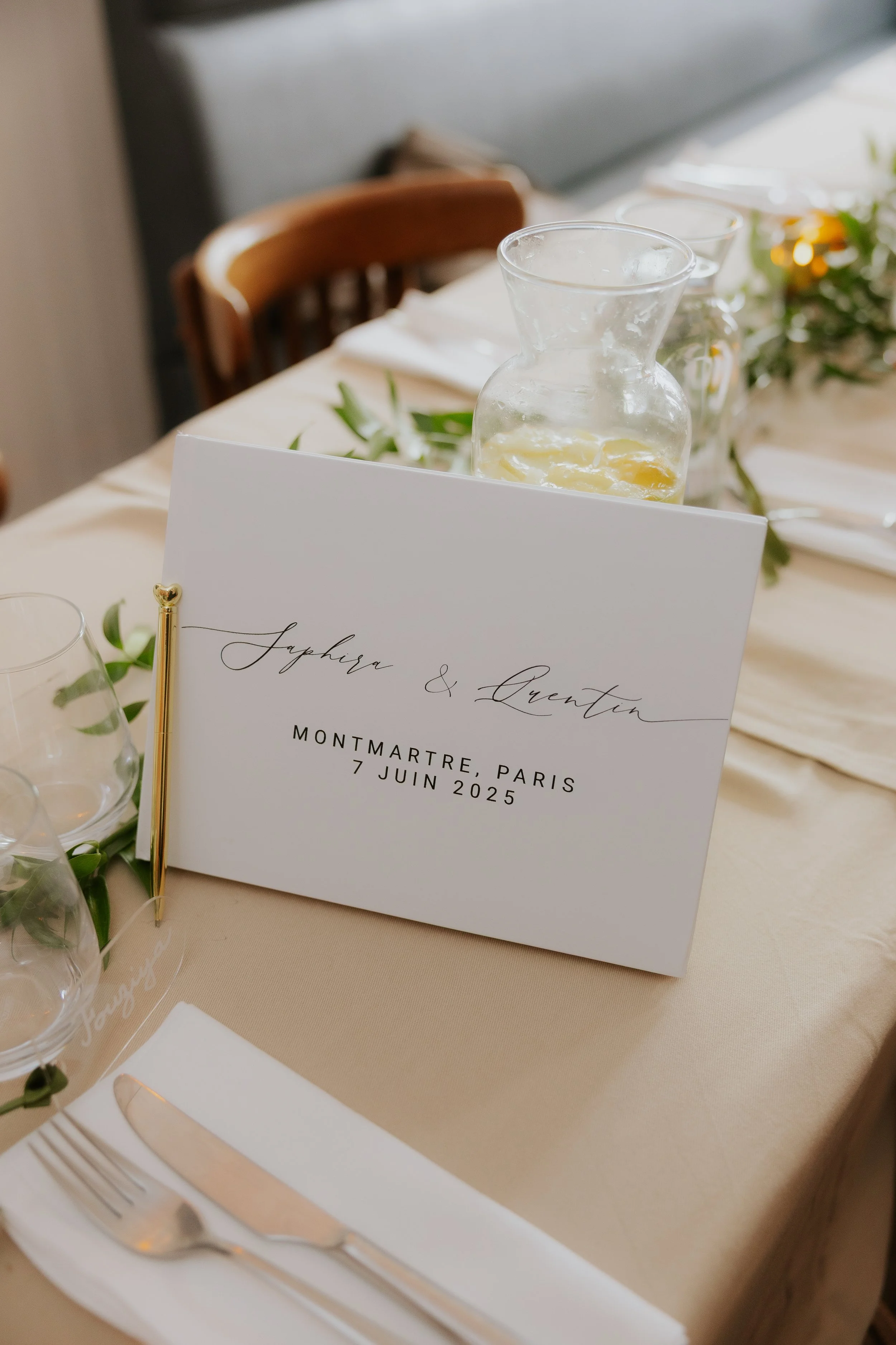 Carte de mariage avec les noms Sapphire et Quentin, indiquant la date et le lieu du mariage à Montmartre, Paris, le 7 juin 2025, placée sur une table de fête avec des verres, des couverts et une carafe d'eau ou de limonade.