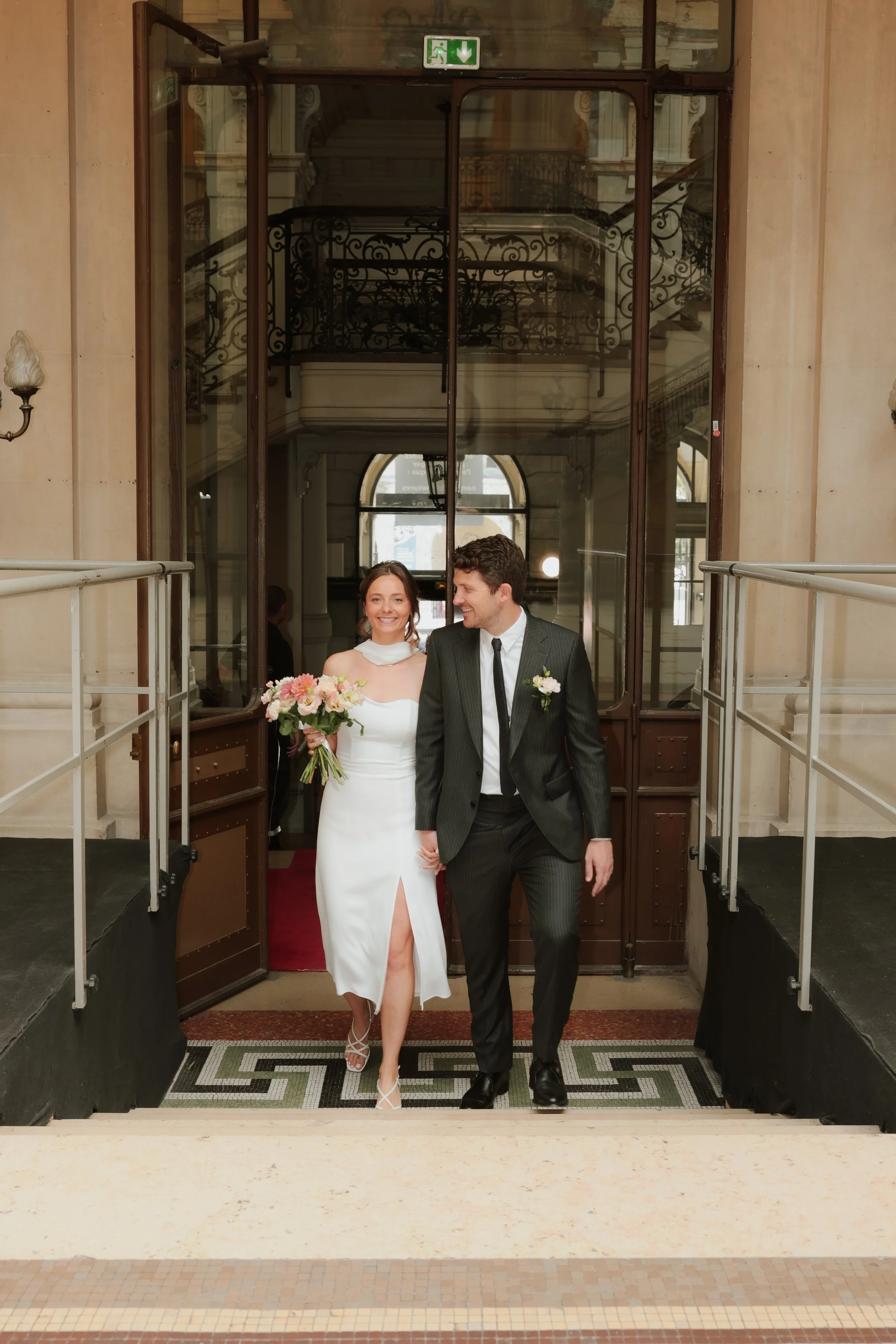 Un couple de mariés qui marche main dans la main à l'entrée d'un bâtiment élégant, la femme tient un bouquet de fleurs et porte une robe blanche, l'homme porte un costume sombre.