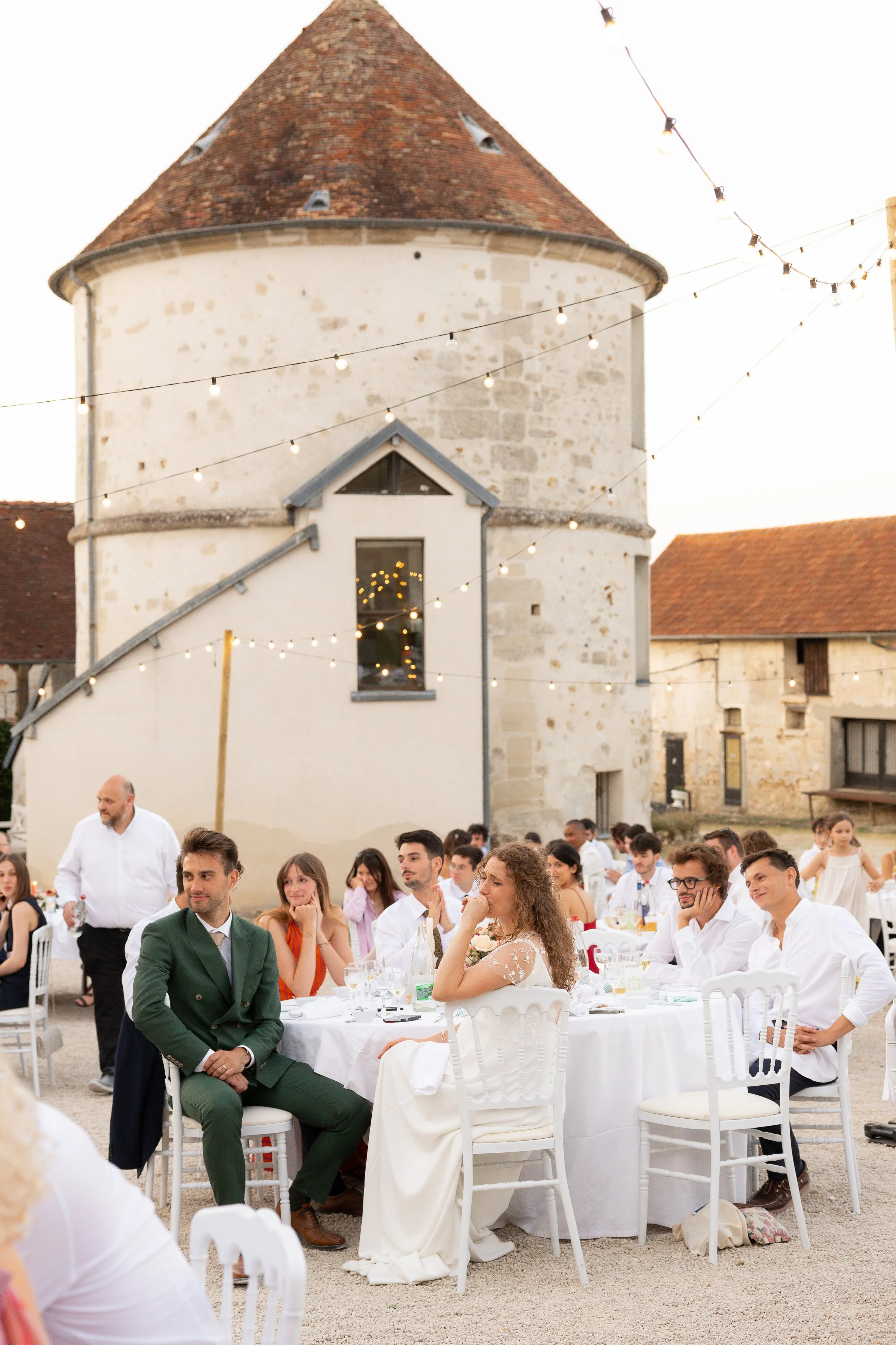 Un groupe de personnes assises autour de tables lors d'un événement en plein air, avec un bâtiment ancien en pierre et un toit en tuiles en arrière-plan. L'ambiance est festive avec des guirlandes lumineuses.