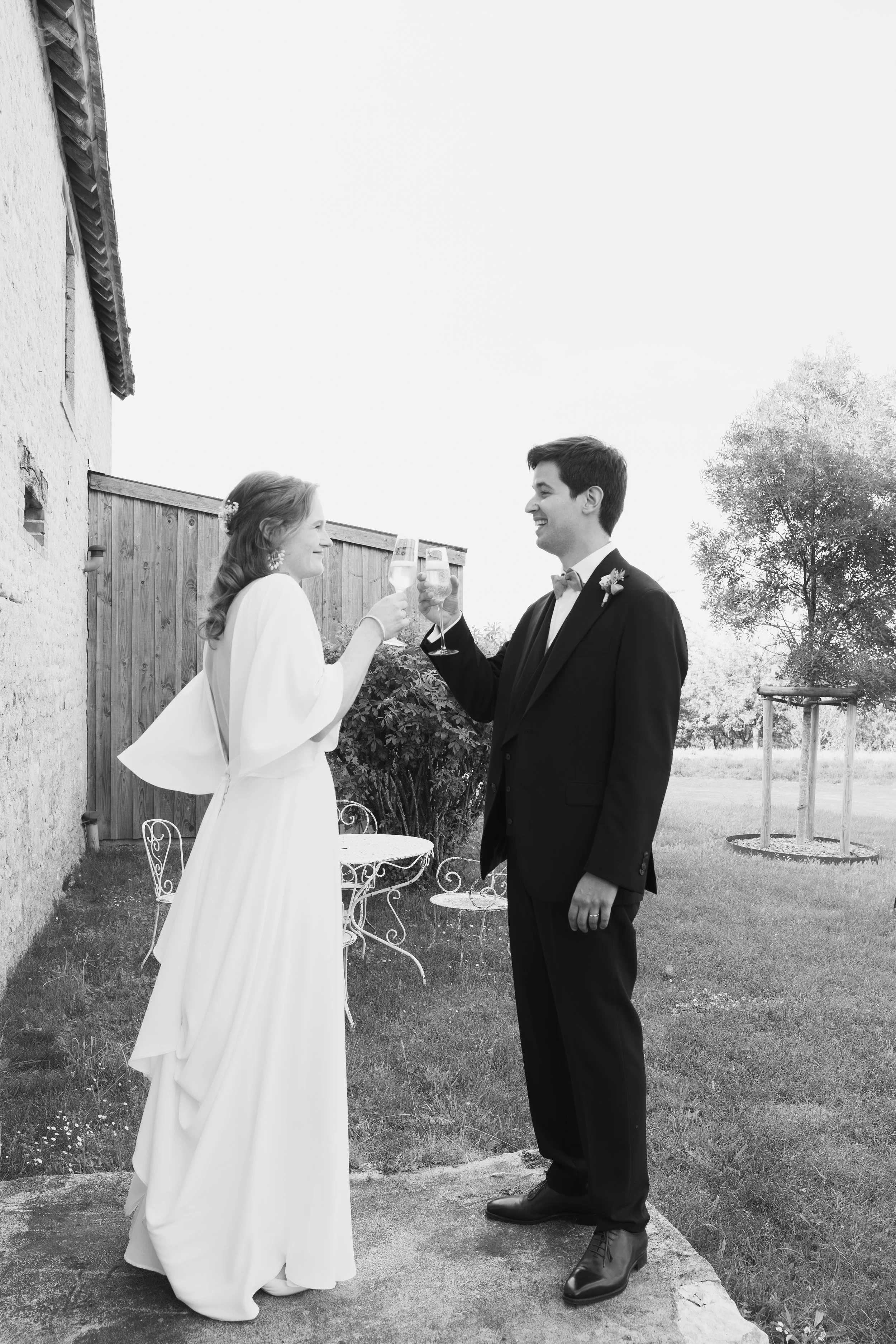 Un couple habillé élégamment échangeant un toast lors d'une célébration en plein air, avec un vieux mur en pierre et un arbre en fond.