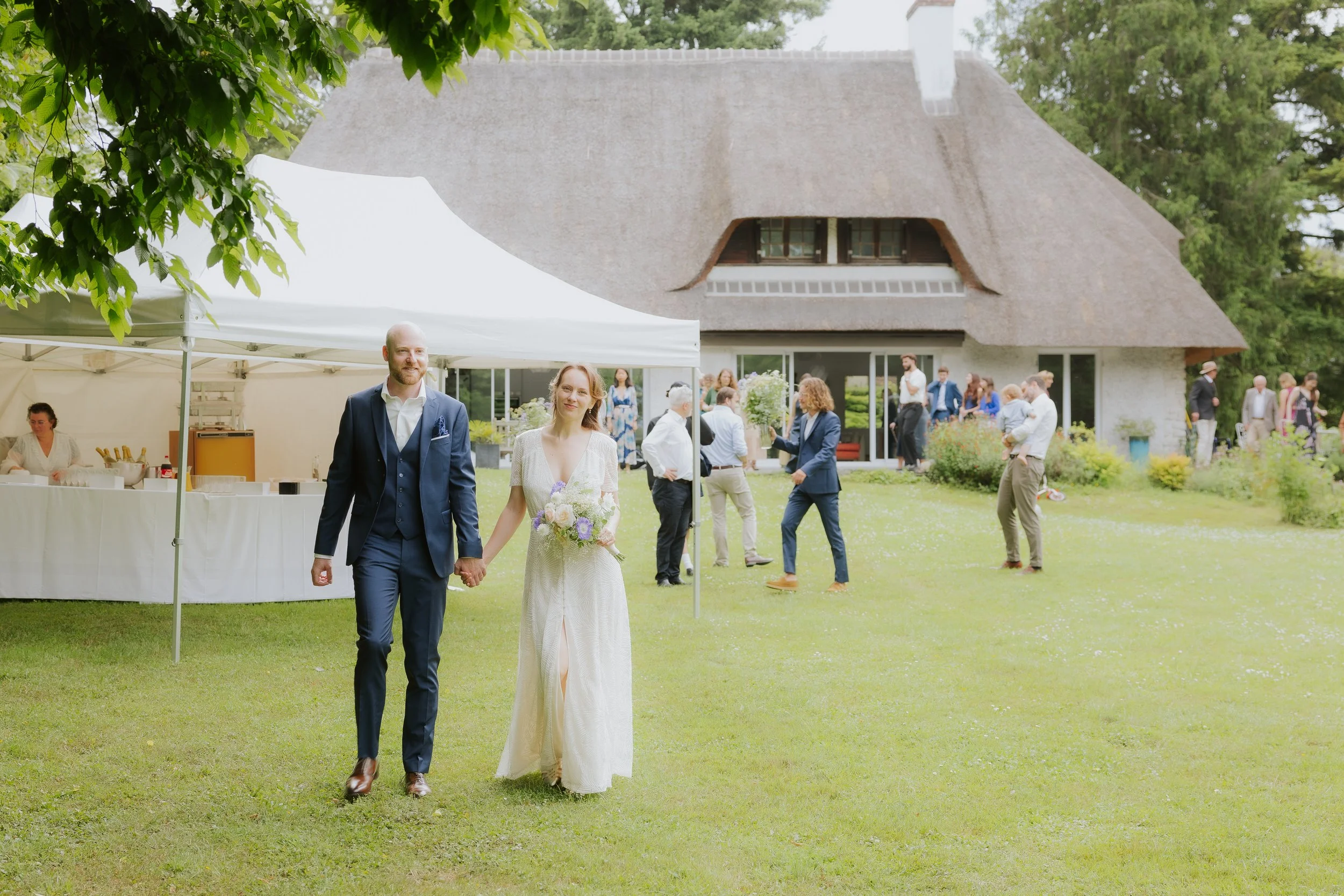 Un couple marié marche main dans la main dans un jardin lors d'une célébration en plein air, avec un groupe de personnes en arrière-plan et une maison à toit de chaume.