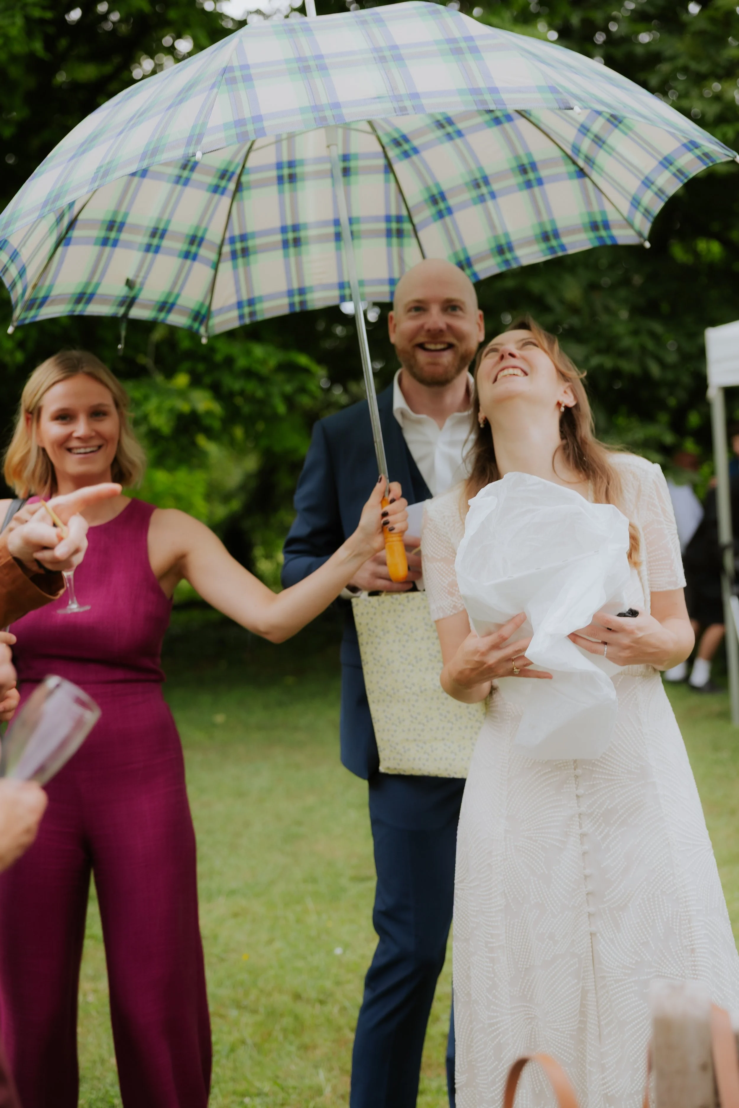 Un groupe de personnes souriantes, dont une femme habillée en blanc, lors d'une célébration en plein air sous un parapluie à carreaux. Ils semblent heureux et font la fête.