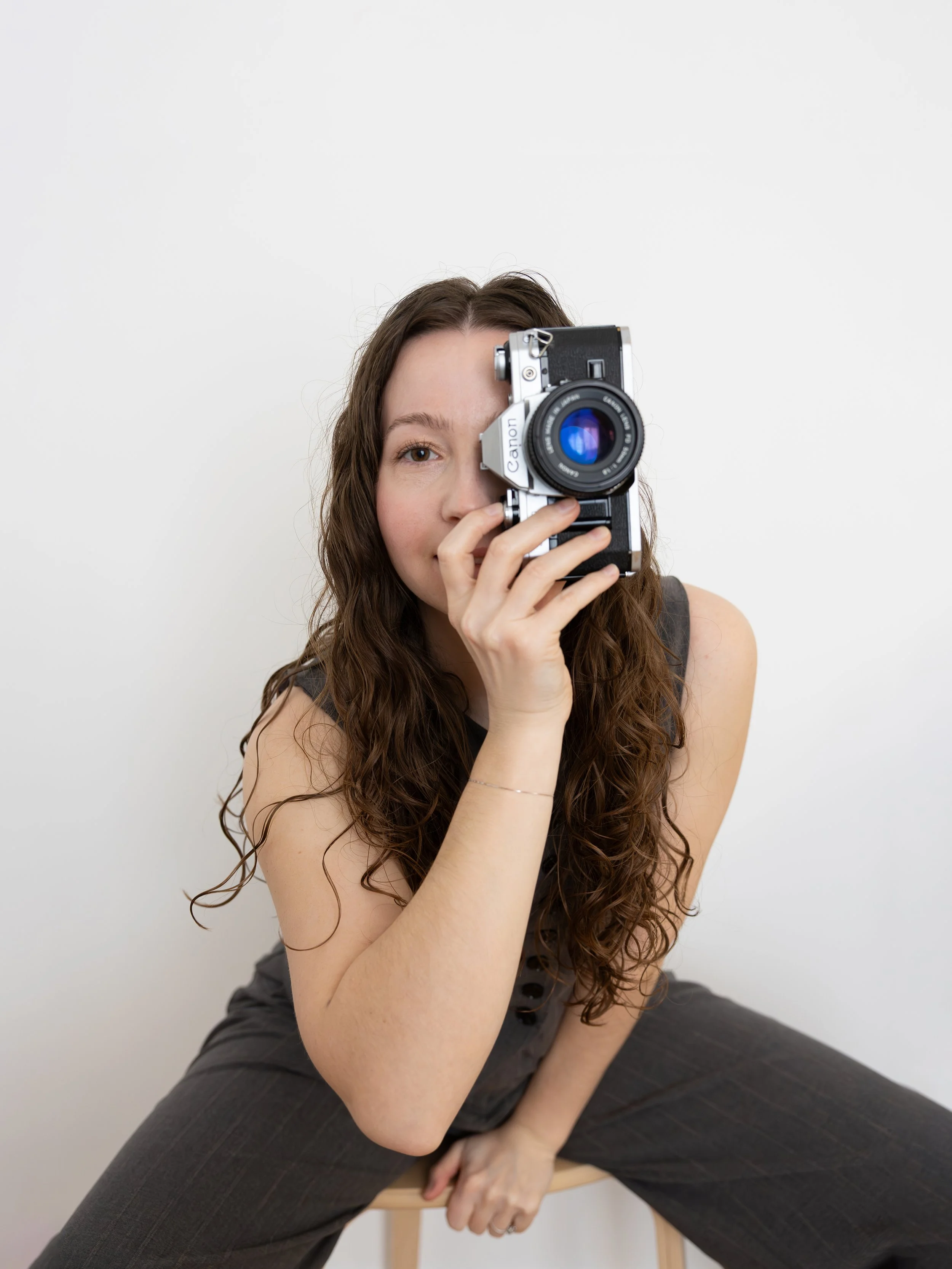 Jeune femme aux cheveux longs, bruns, assise sur une chaise en bois, tenant un appareil photo Canon face à elle, avec un fond blanc.