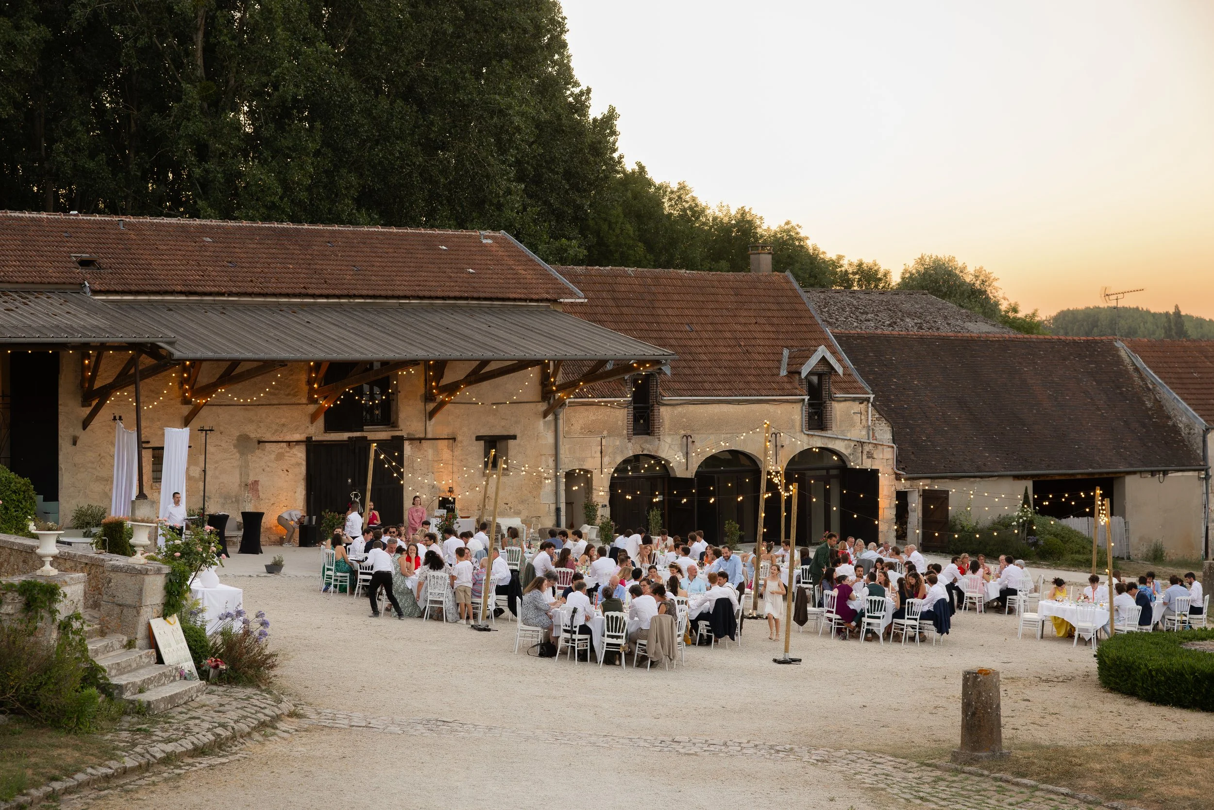 Une grande fête en soirée avec de nombreuses personnes assises à des tables à l'extérieur devant une vieille bâtisse en pierre avec toits en tuiles rouges, décorée de guirlandes lumineuses, dans un cadre rural.