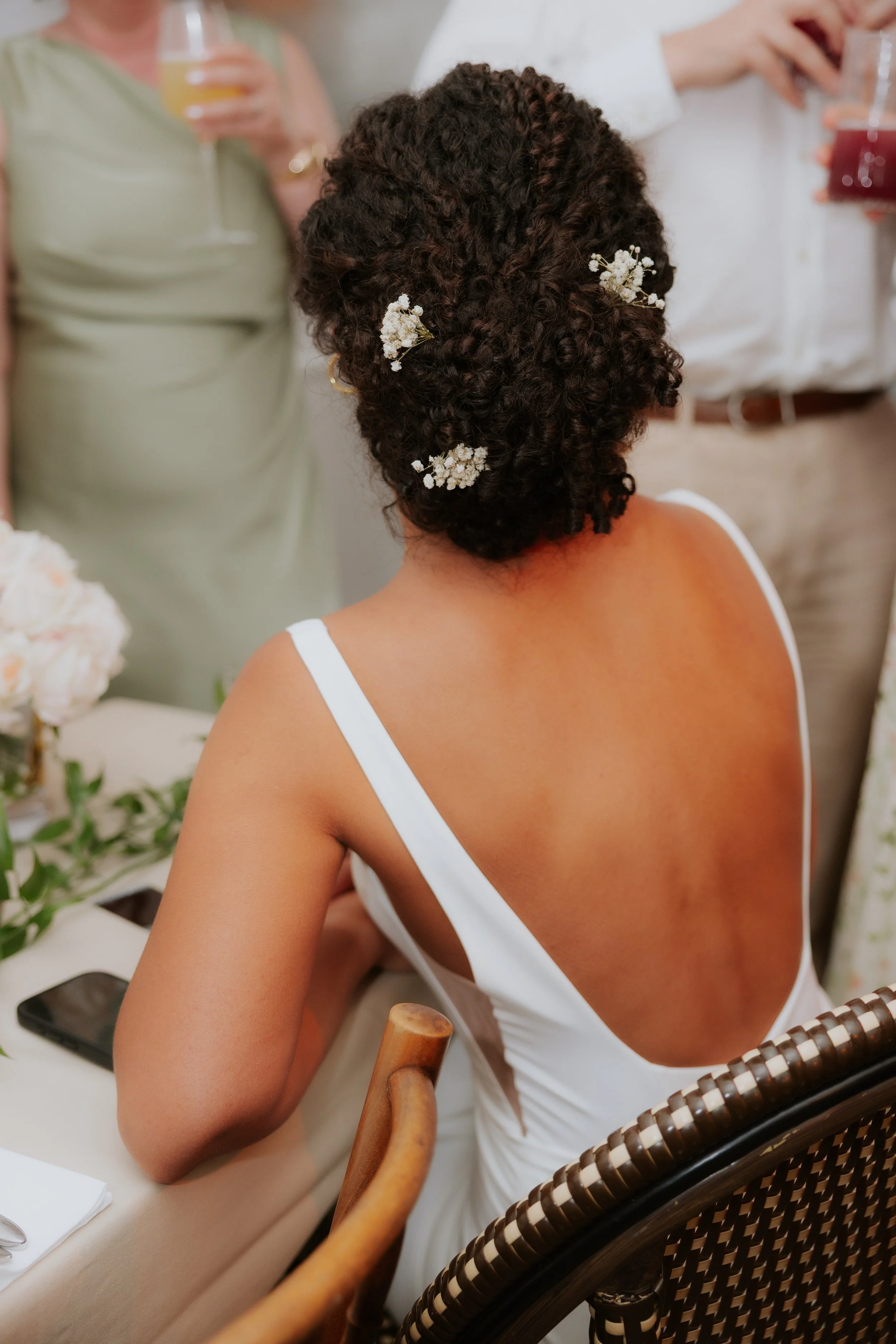 Femme avec cheveux bouclés décorés de petites fleurs, en robe blanche avec dos dénudé, assise à une table lors d'une célébration ou fête.