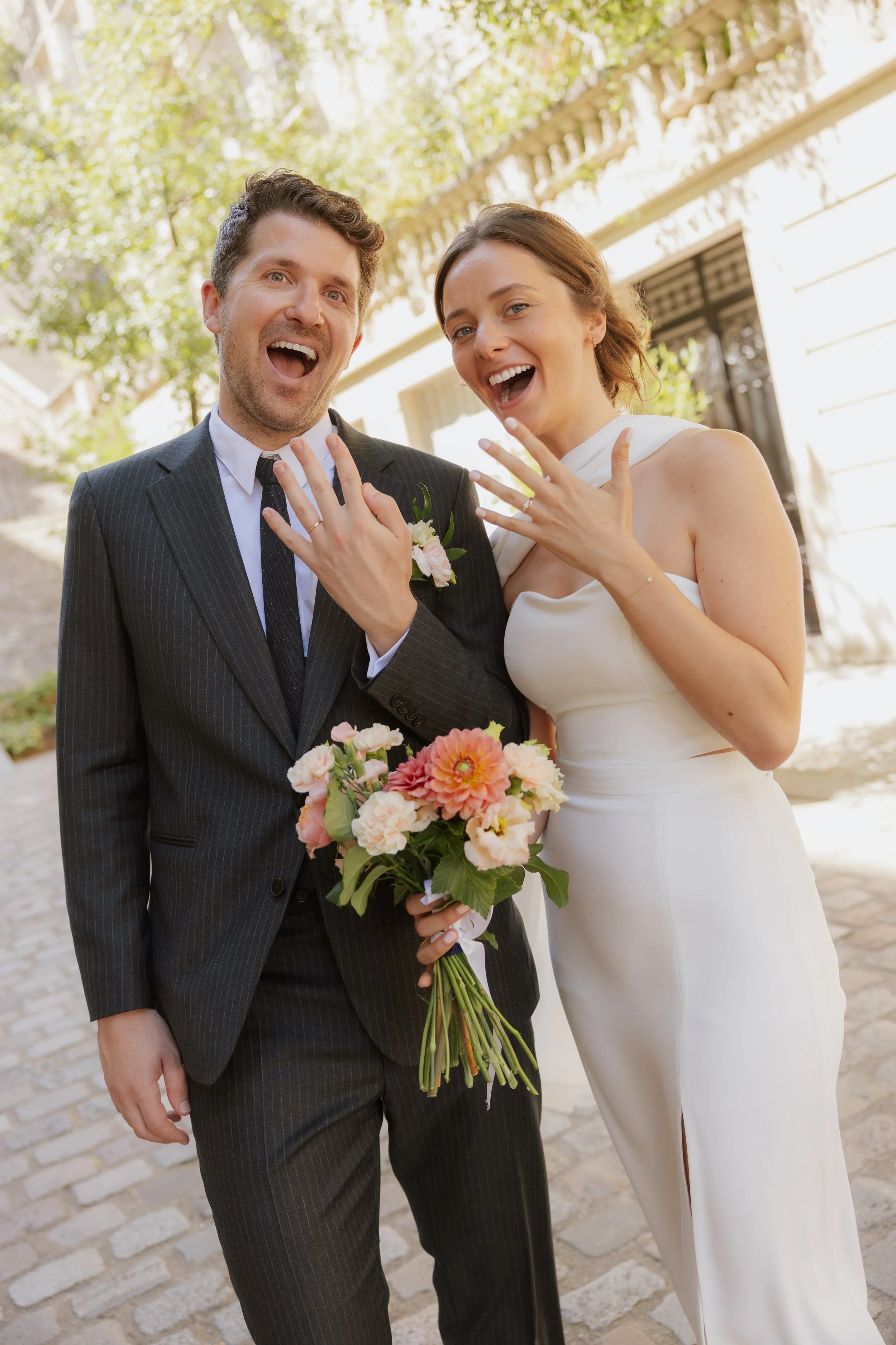Un couple de mariés souriants montrant leurs alliances, la femme tenant un bouquet de fleurs, devant une maison en extérieur.
