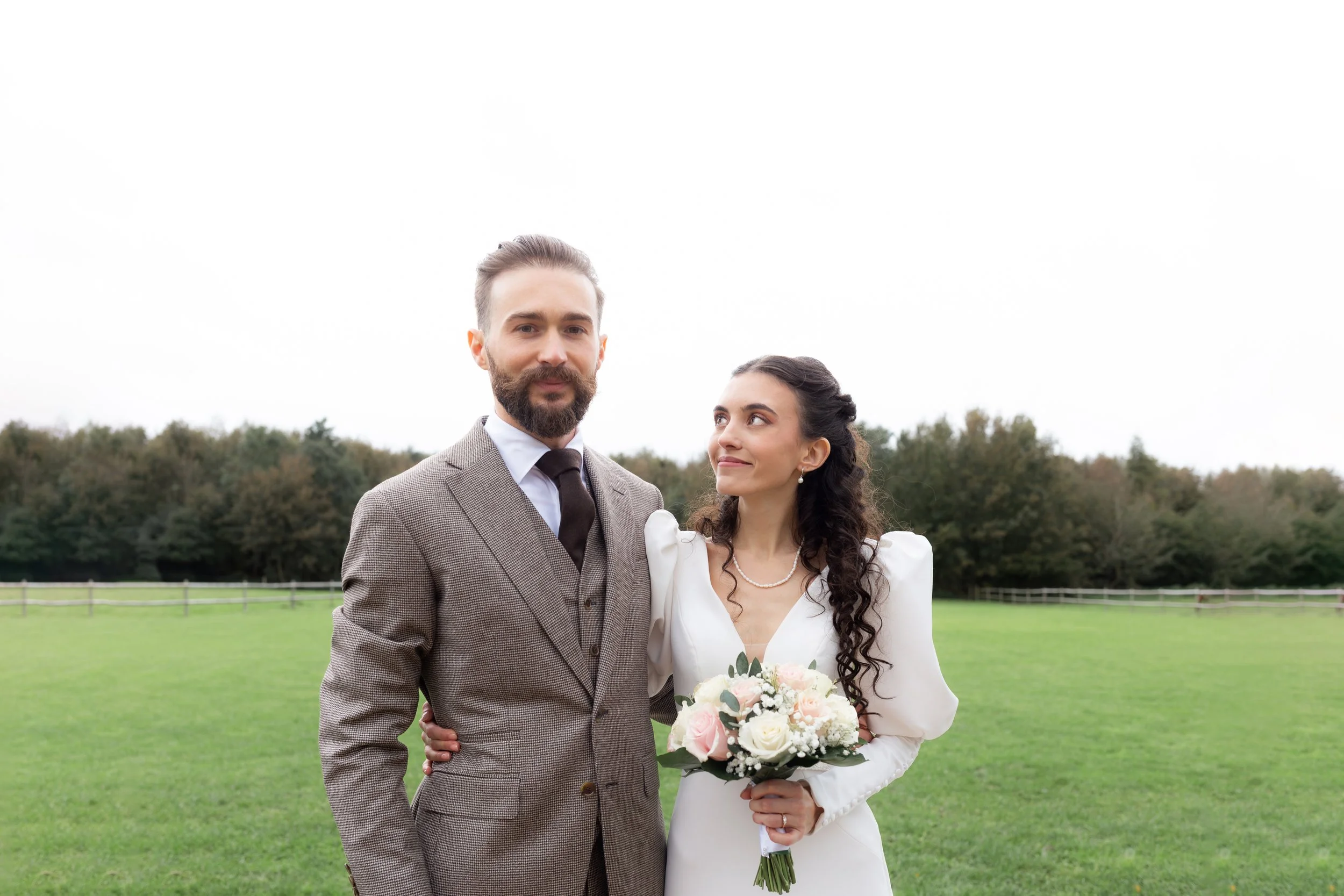 Un couple en costume de mariage pose dans un champ vert, la femme tient un bouquet de fleurs blanches et roses, en arrière-plan des arbres et un ciel nuageux.