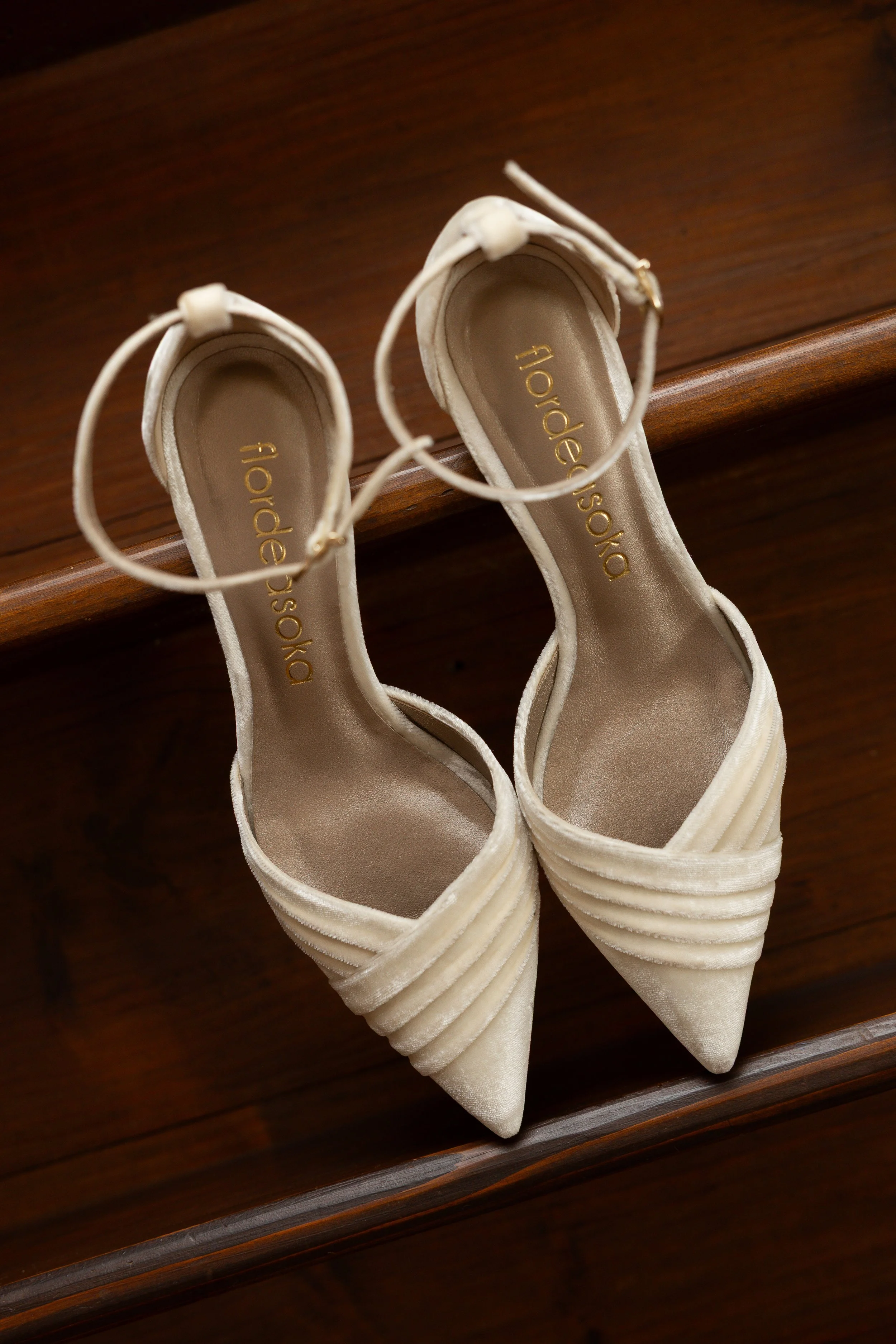 Une paire de chaussures de mariée beige en satin avec des talons hauts, posées sur une étagère en bois.