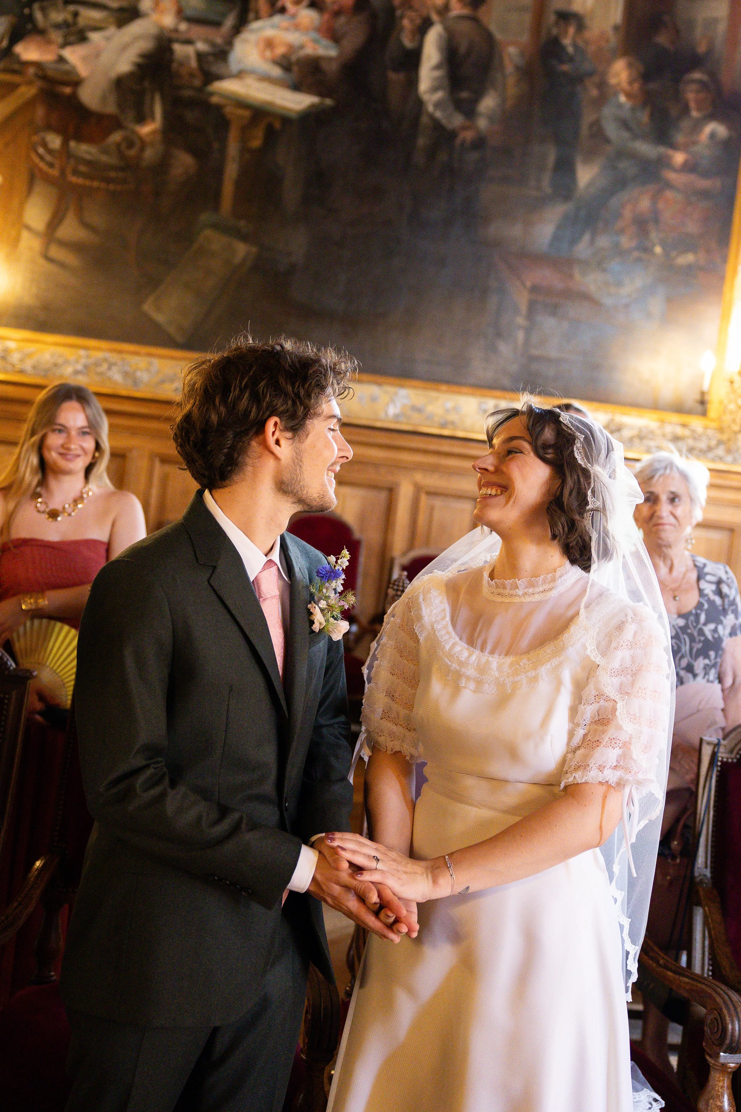 Un couple en train de se marier, s'tenant les mains et se regardant avec bonheur lors de la cérémonie. Autres personnes visibles en arrière-plan, dans un lieu intérieur décoré élégamment.