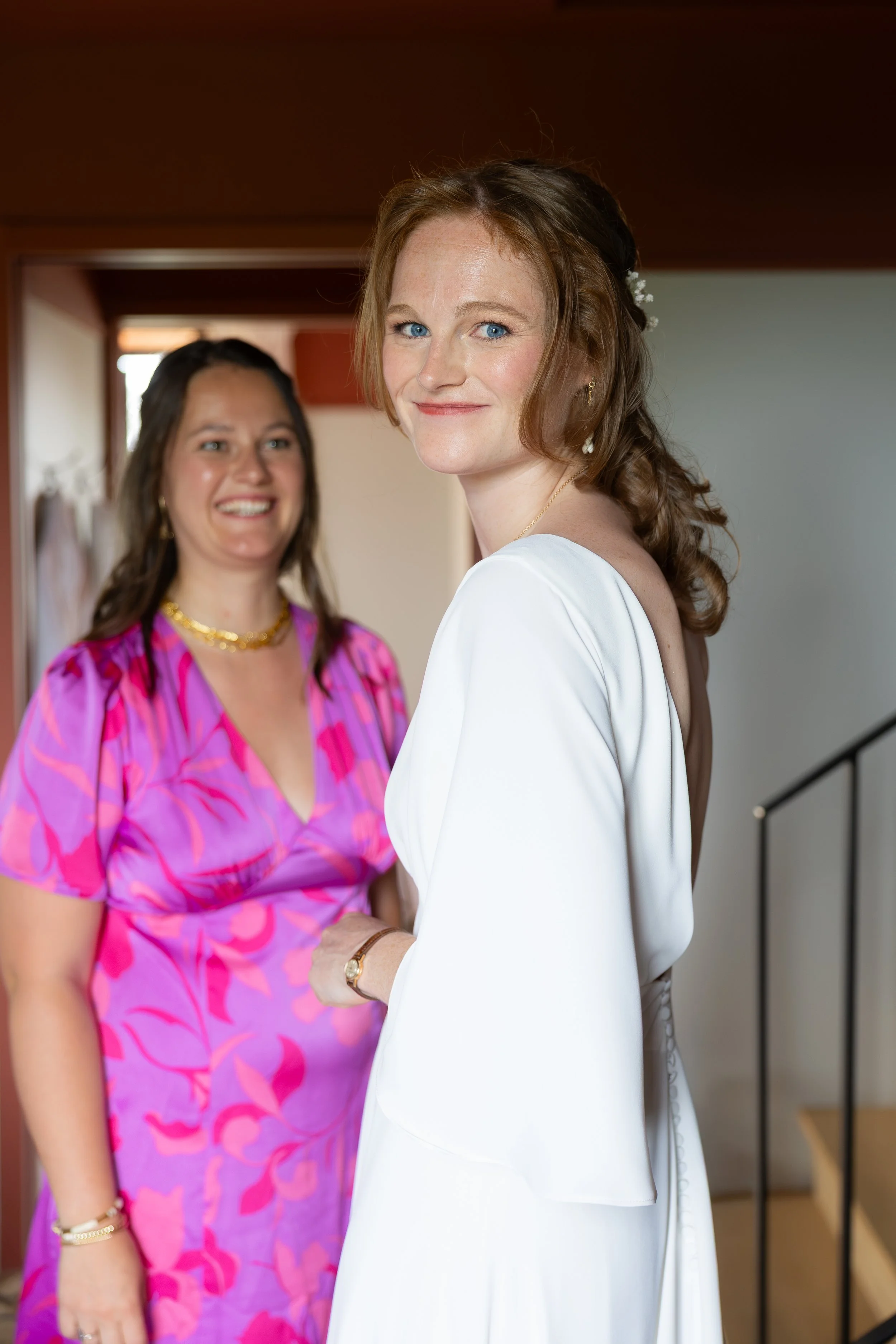 Deux femmes, l'une en robe blanche et l'autre en robe mignonne à motifs roses et violets, sourient dans un intérieur lumineux.